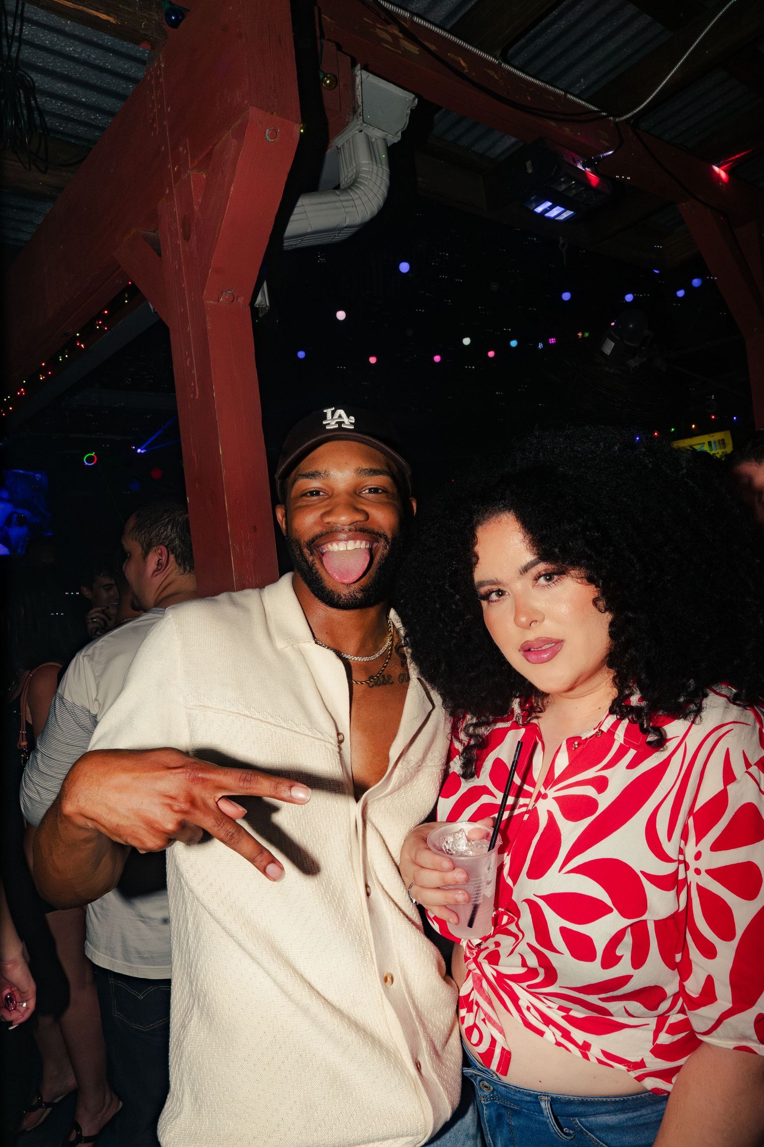 Two friends smiling and posing together at a nightclub, man in LA cap and woman in red floral top.