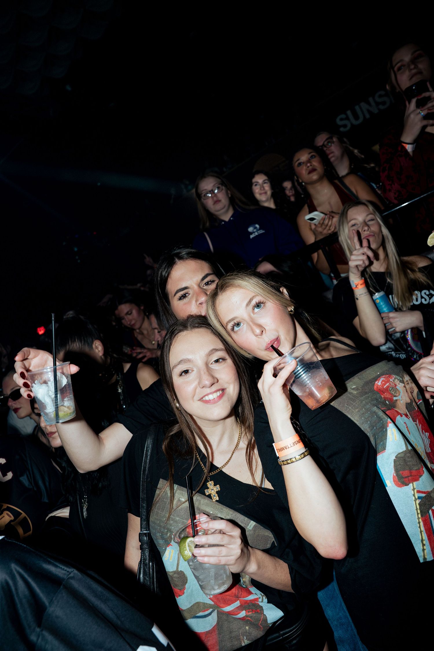 Two young women smiling and drinking at a crowded concert venue at night.