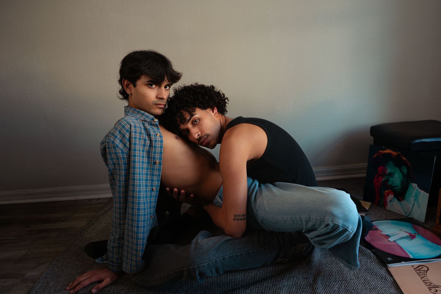 Two young people sit closely together on a floor, one resting their head on the other.