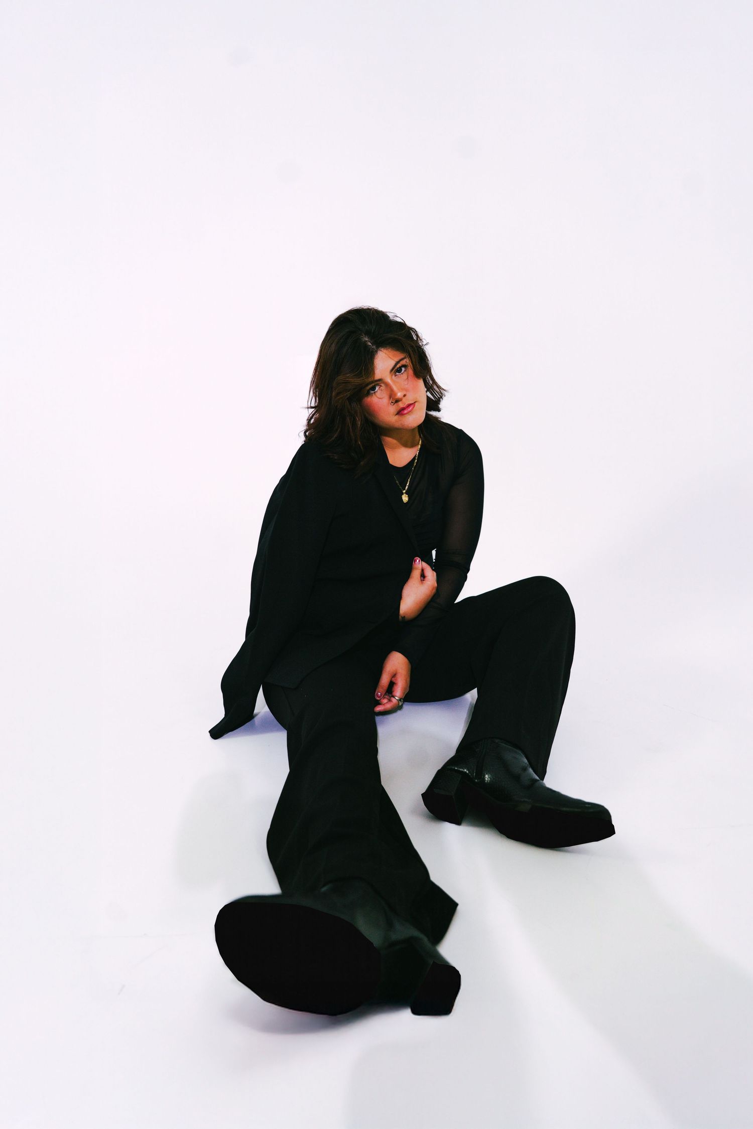 Young woman in all-black outfit sitting on white studio floor wearing chunky black boots posing.