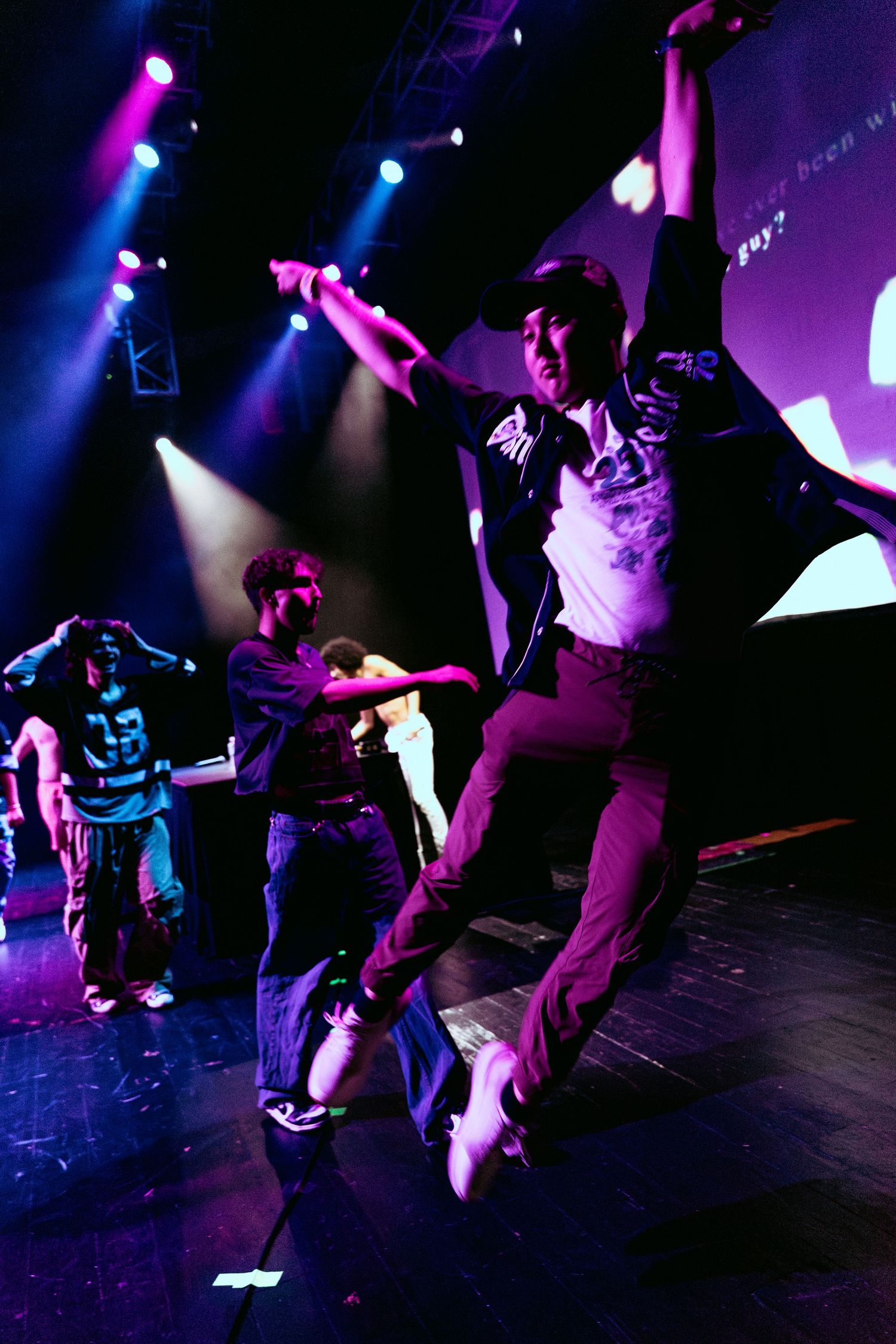 A performer leaps energetically on stage under colorful purple and blue stage lights during a live dance show.