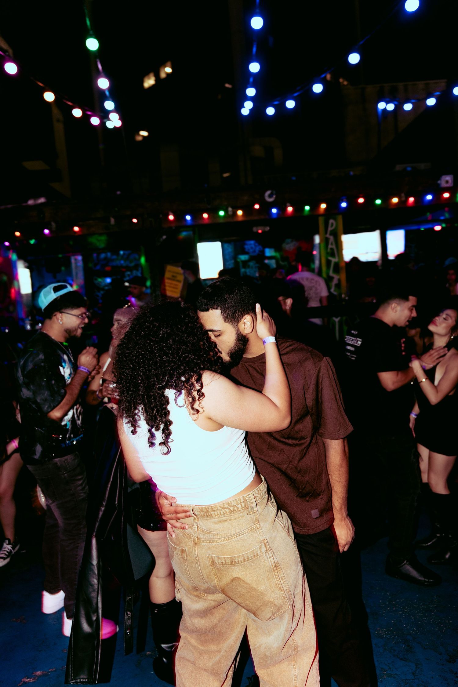 Couple sharing a romantic moment on a crowded dance floor at a vibrant nightclub with colorful lights.