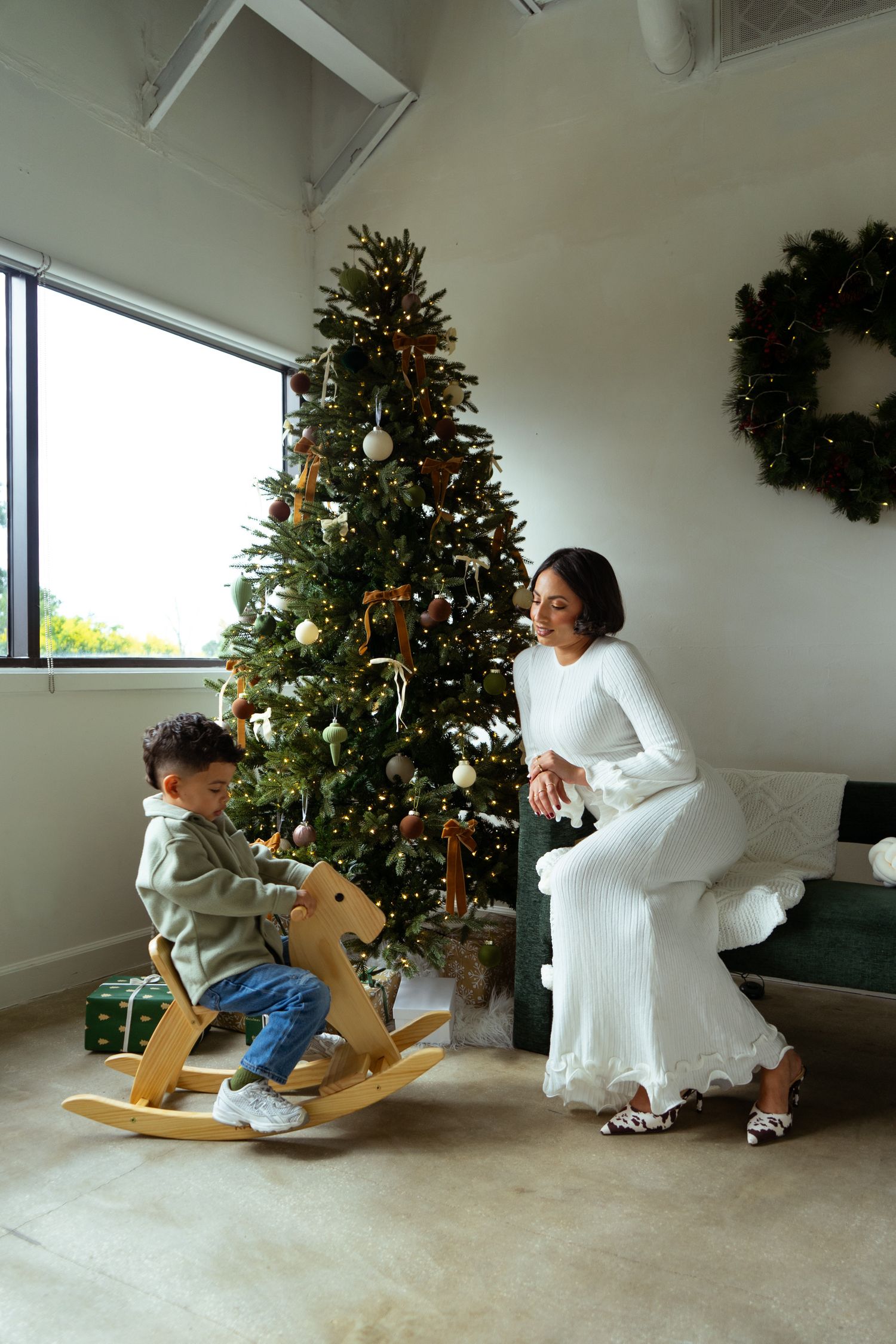 Woman in white dress watching a young boy ride a wooden rocking horse near a decorated Christmas tree.