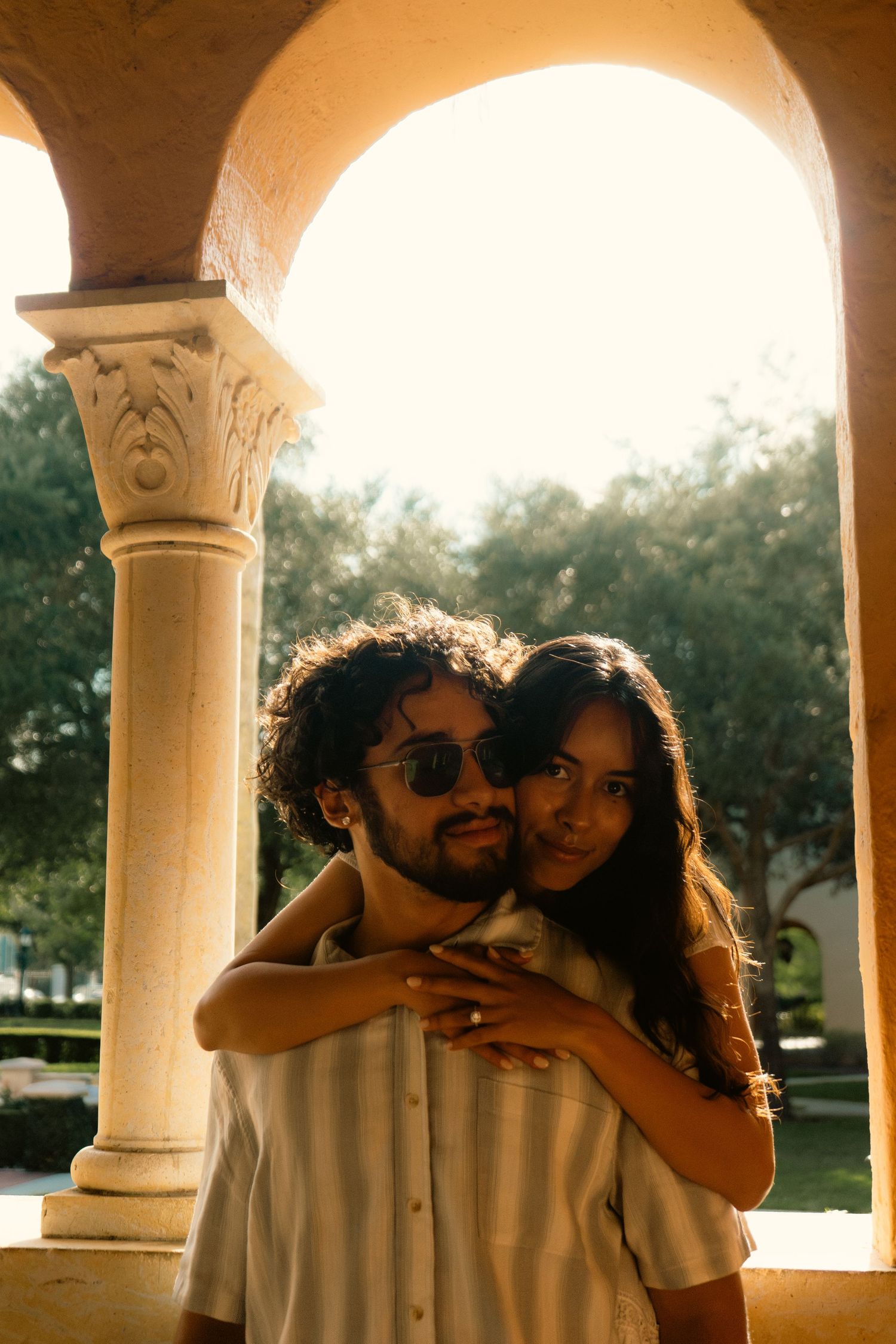 Couple embracing under ornate stone arch with column in warm golden sunlight outdoors.
