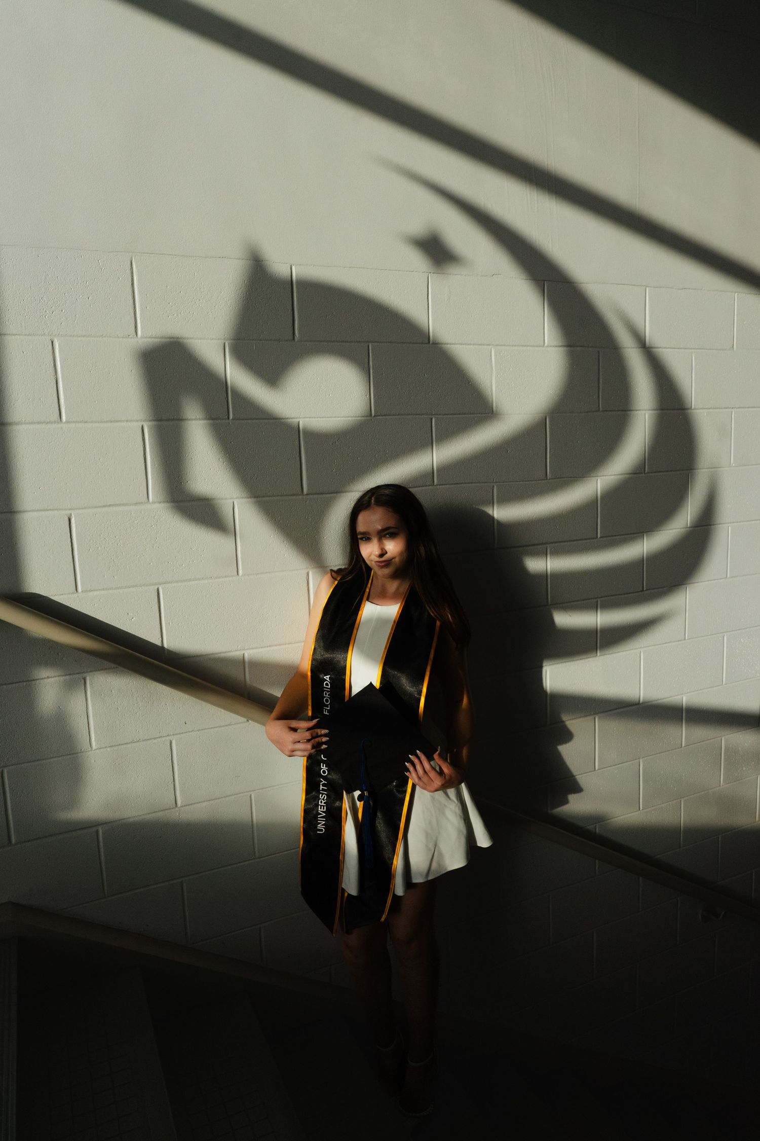 Graduate in black and gold sash poses by wall with university logo shadow at commencement ceremony.