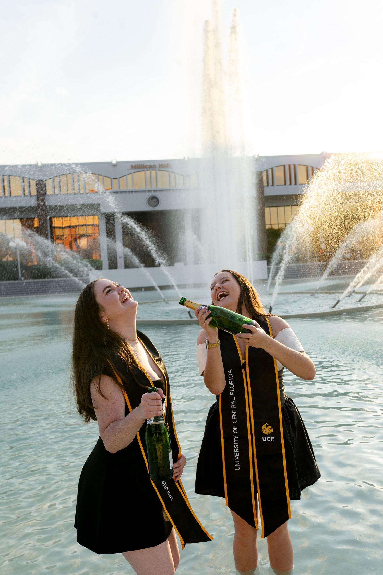 Two UCF graduates laughing and popping champagne bottles by a fountain on campus.