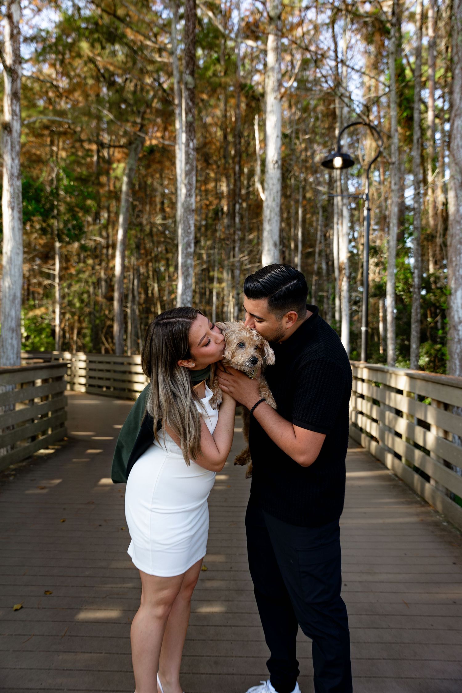 Couple kissing their small dog on a wooden boardwalk surrounded by tall trees.