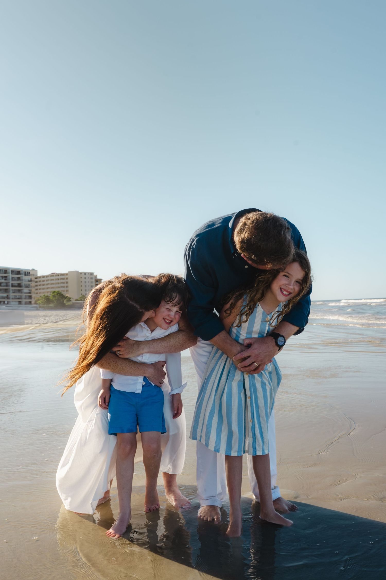 Family laughing and embracing together on a sunny beach with clear blue skies.