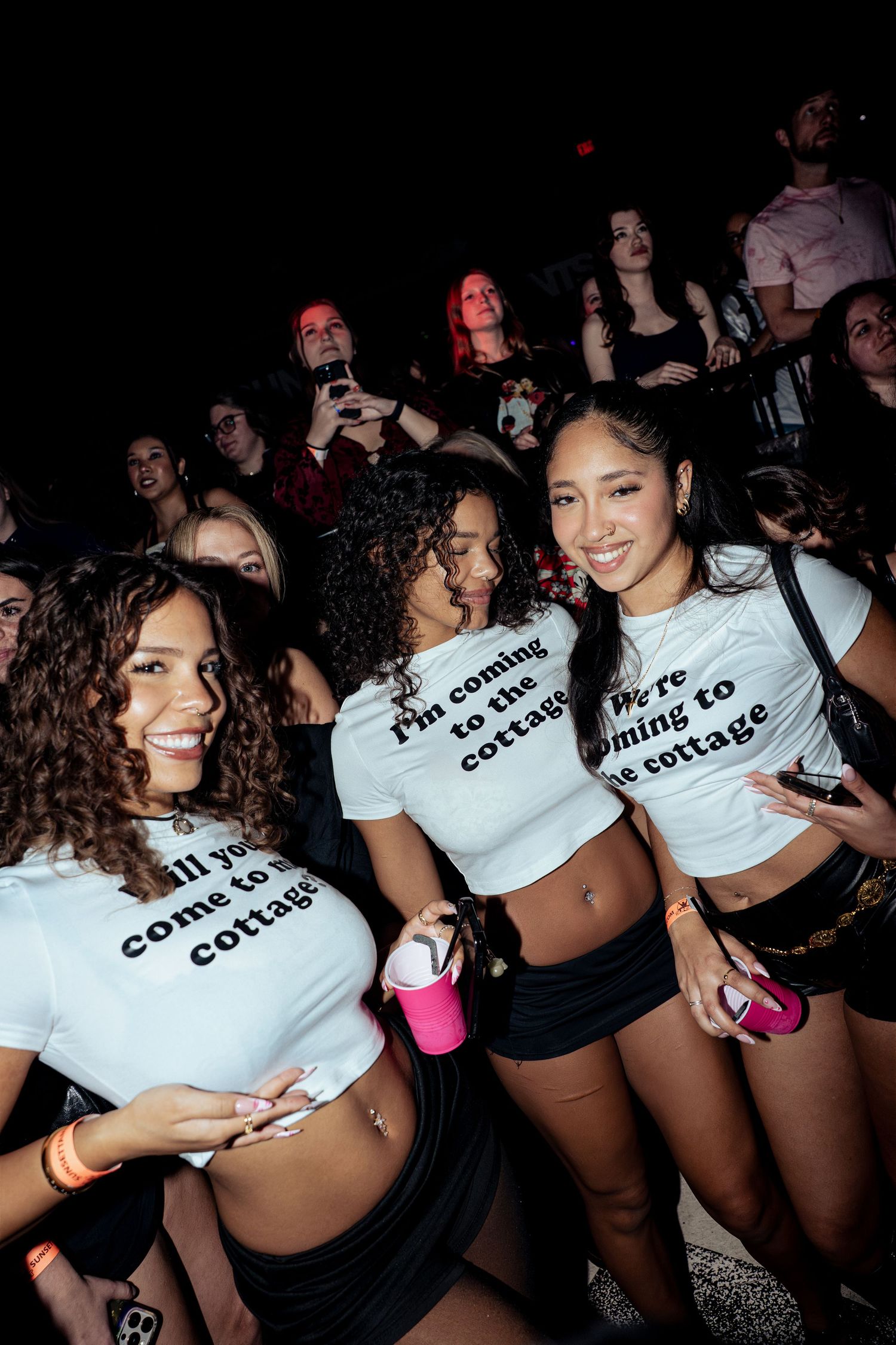 Three smiling women in matching white crop tops at a nightclub event posing together.