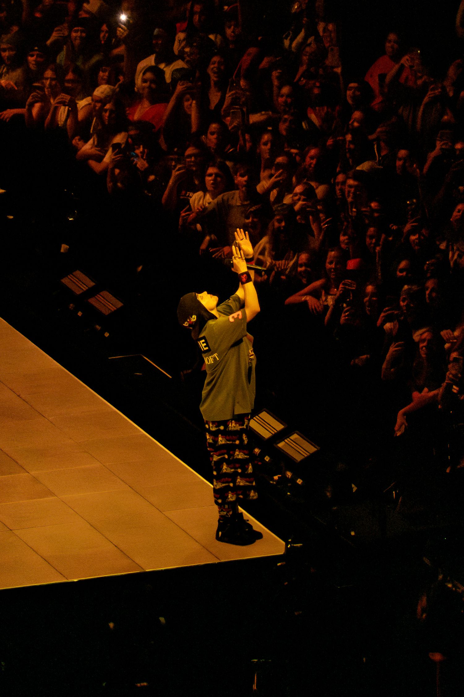 Concert performer raises hands to crowd in a large arena illuminated by warm golden light.
