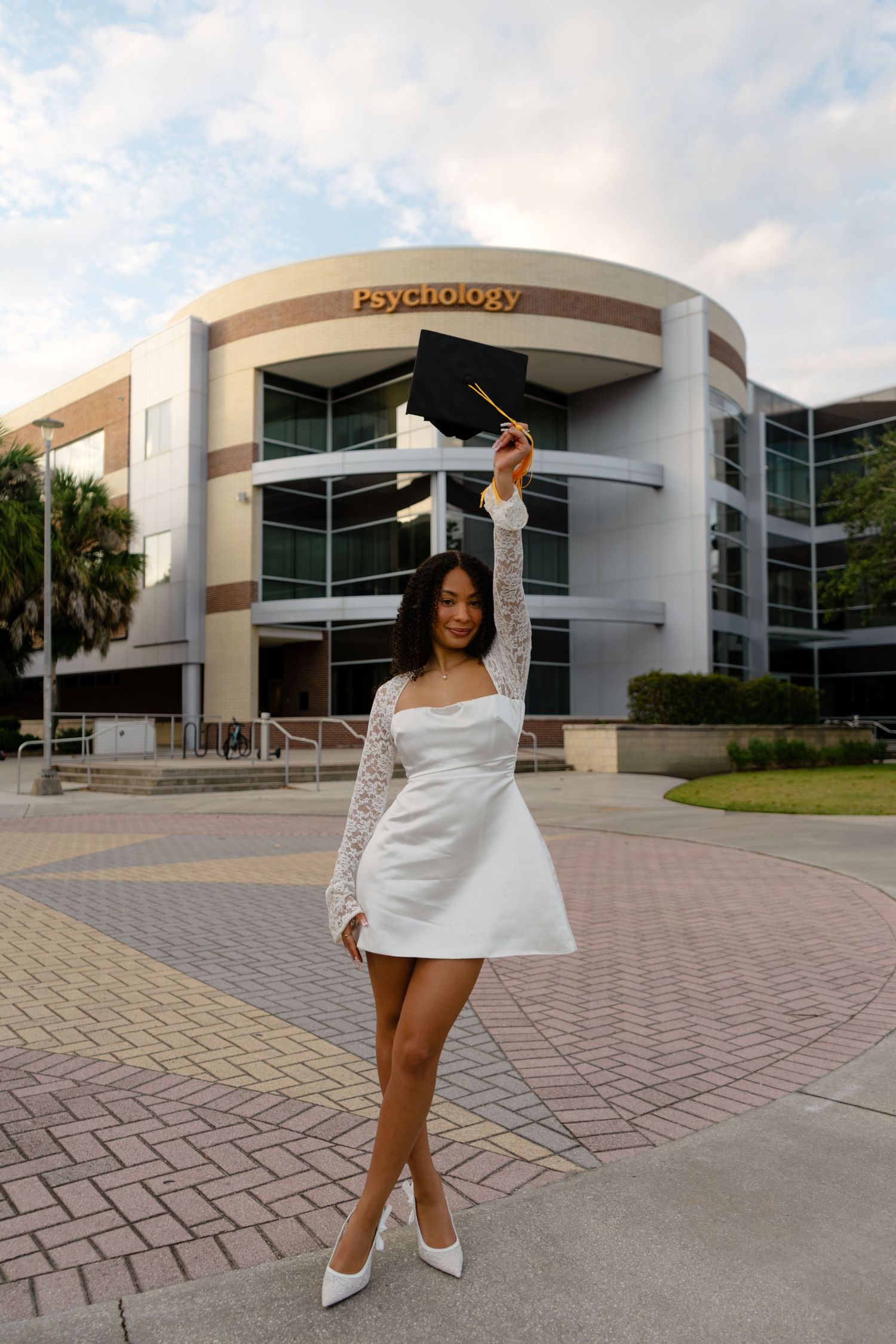 Graduate in white dress raises cap triumphantly in front of Psychology building on university campus.