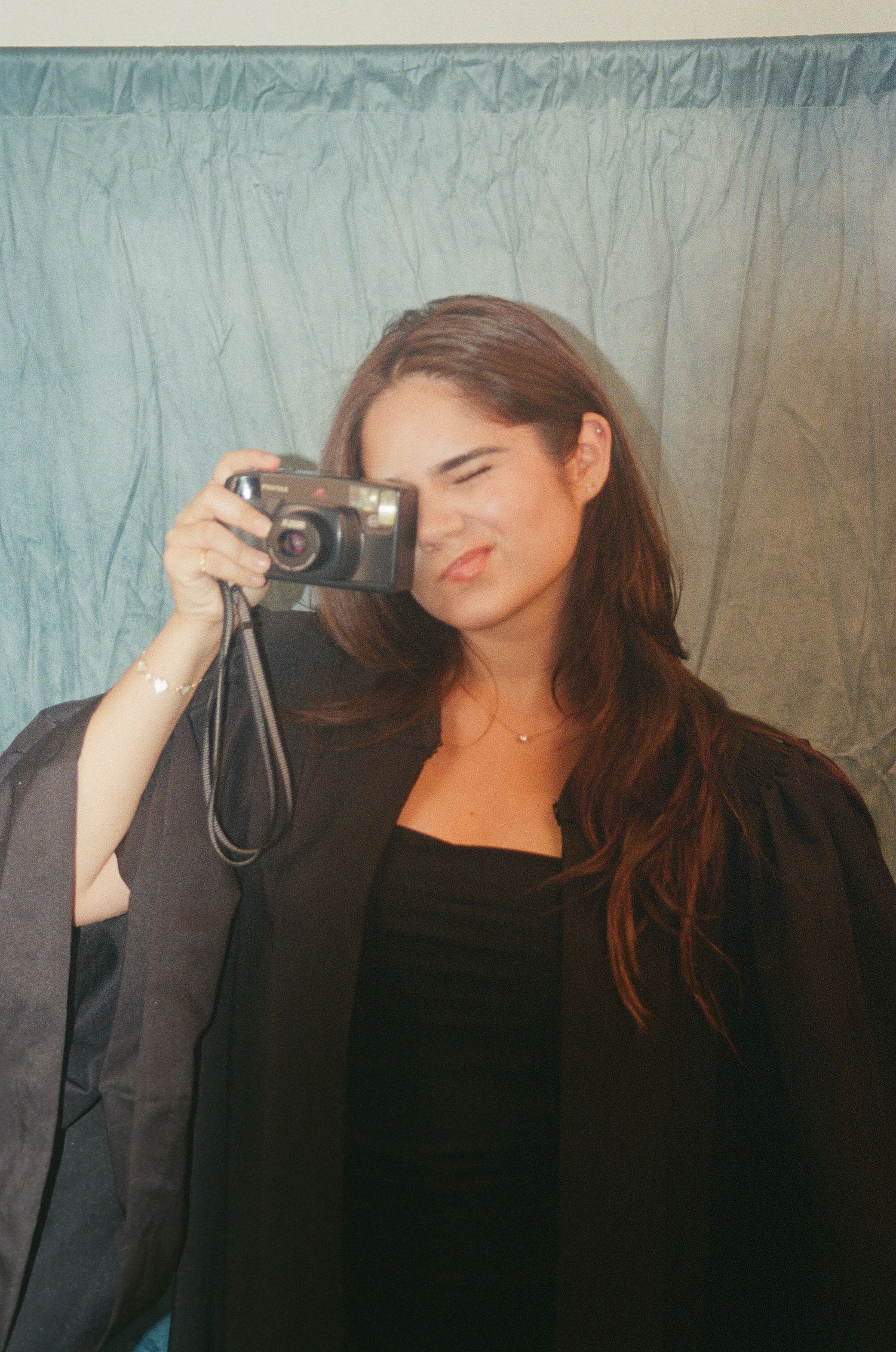 Graduate in black gown holds film camera up to face in mirror selfie.