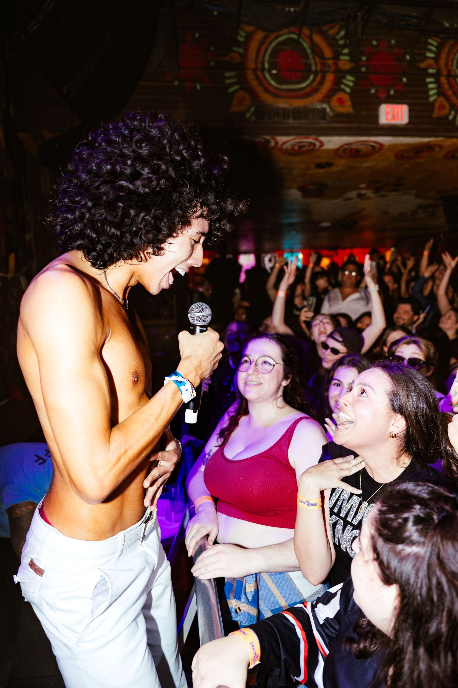 Shirtless curly-haired performer sings into microphone while engaging with excited crowd at a lively concert venue.