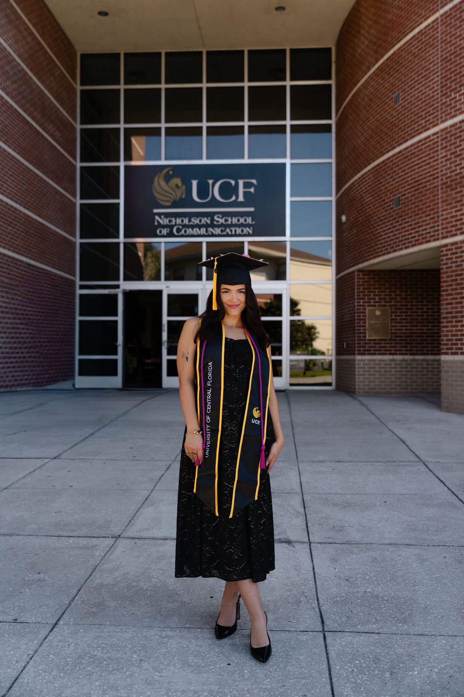Female graduate in cap and gown stands before UCF Nicholson School of Communication building.