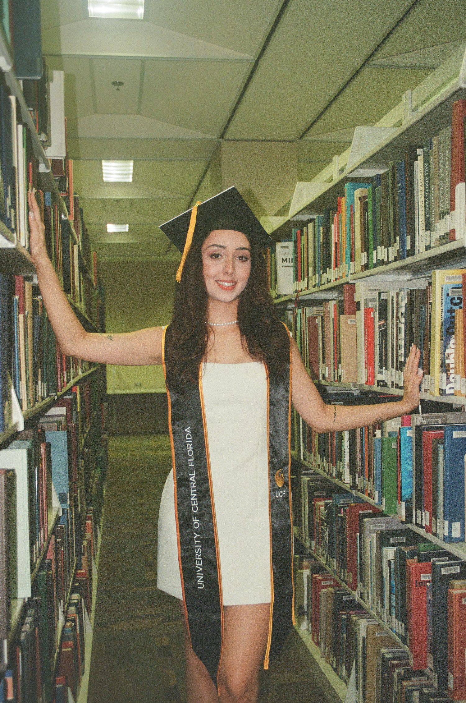 Female UCF graduate in cap and gown poses between library bookshelves in white dress.