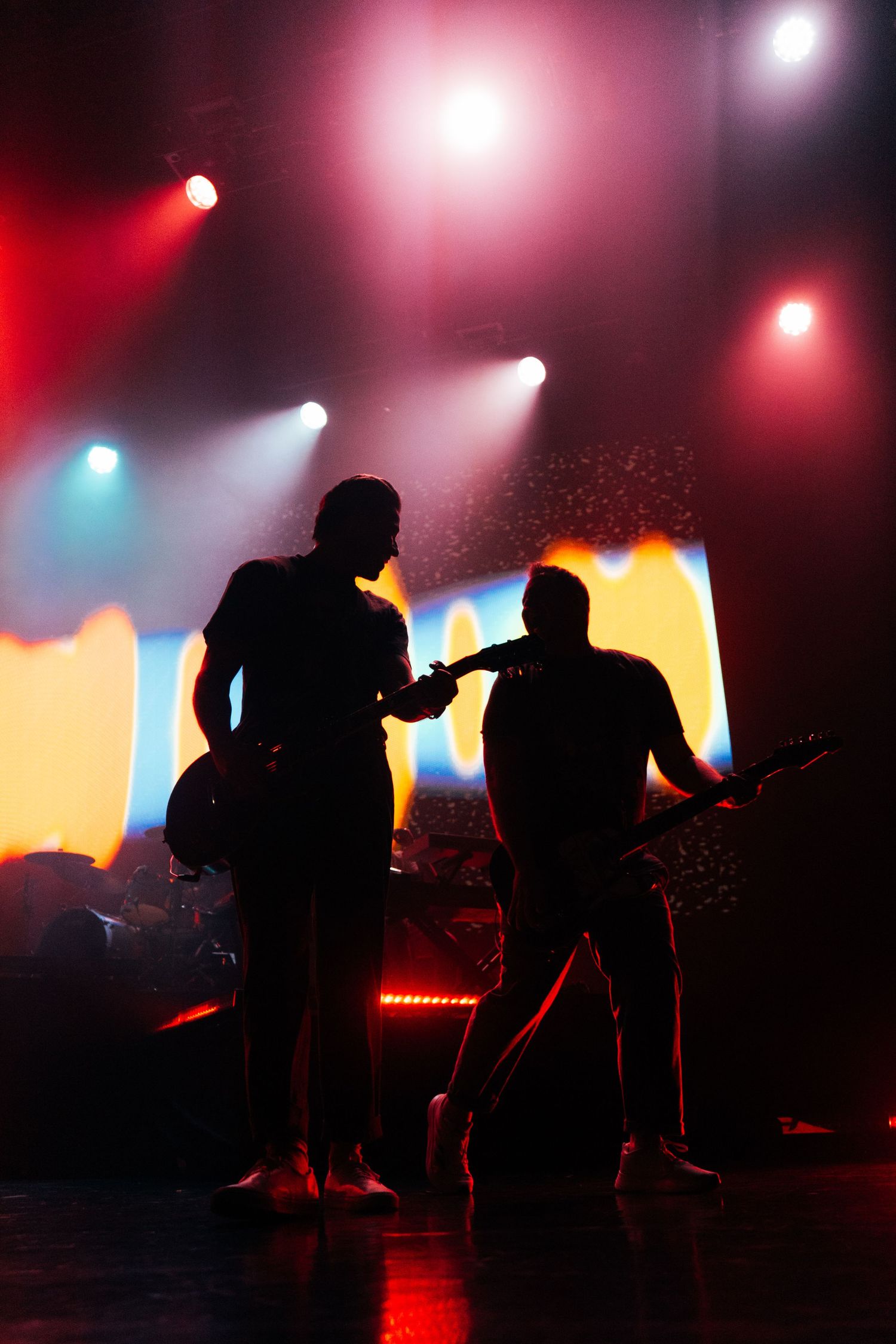 Two guitarists performing on stage as dramatic silhouettes against vibrant red and blue concert lights.