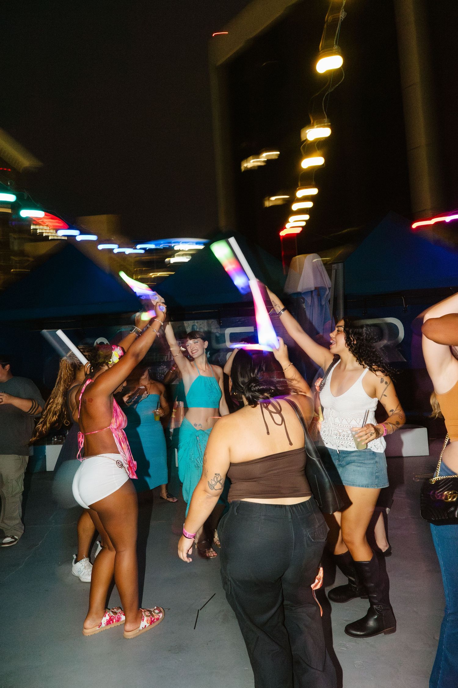 People dancing at an outdoor nightclub event with glowing LED light sticks under blue canopies at night.