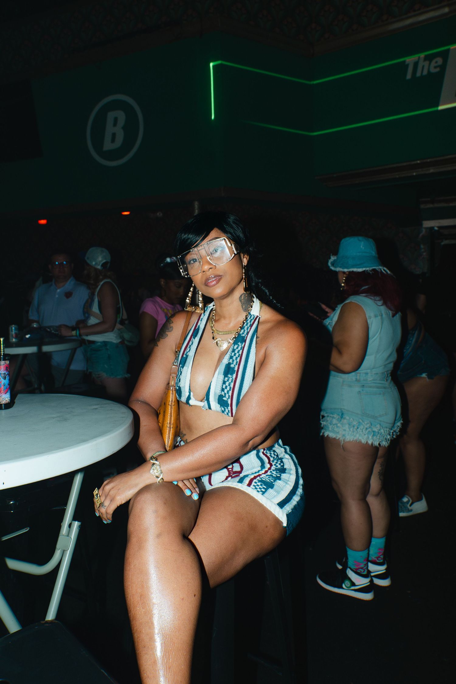 Woman in printed romper posing at a nightclub table under green neon lights with crowd in background.