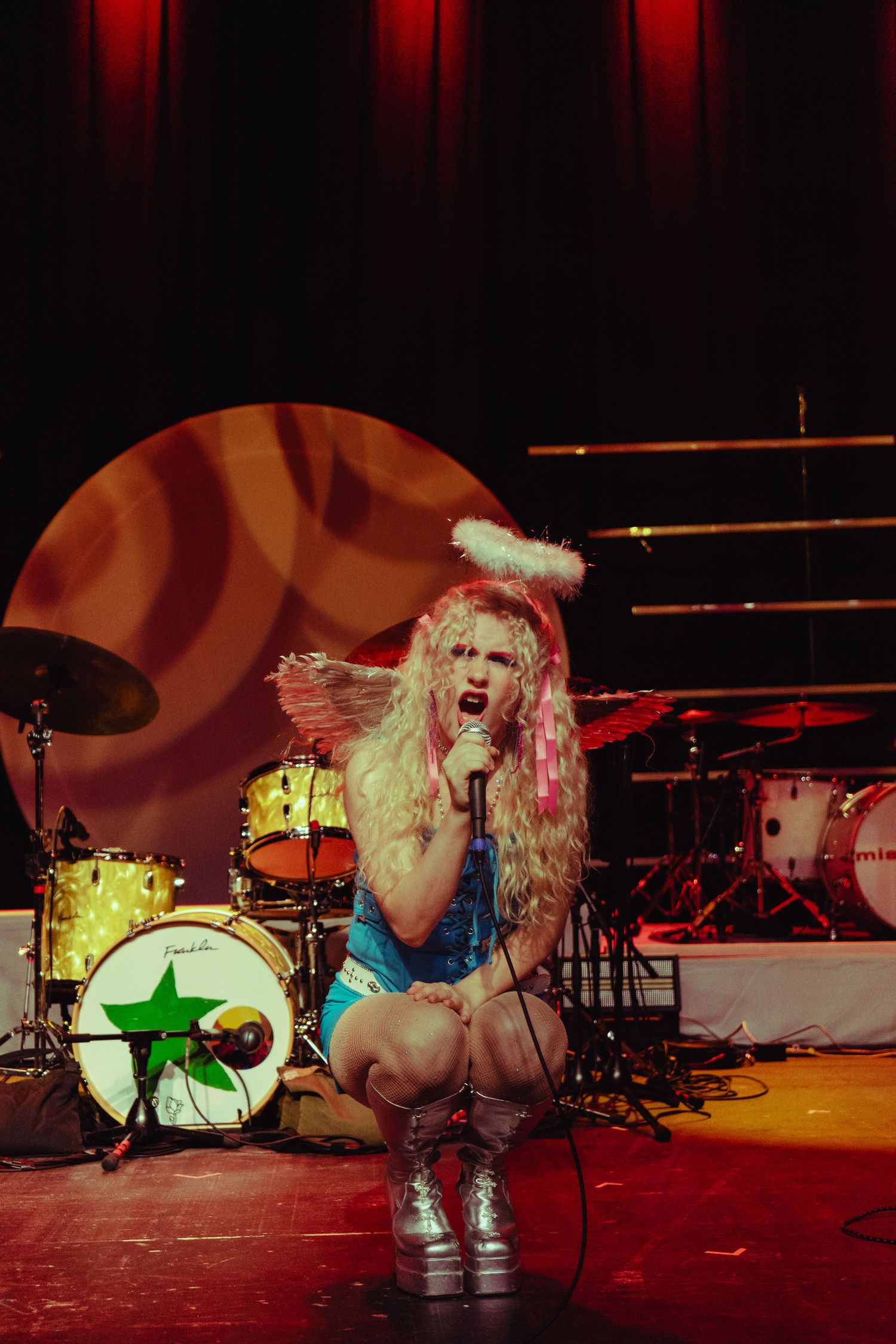 Blonde female performer in blue angel costume kneels on stage with feather wings and halo headpiece.