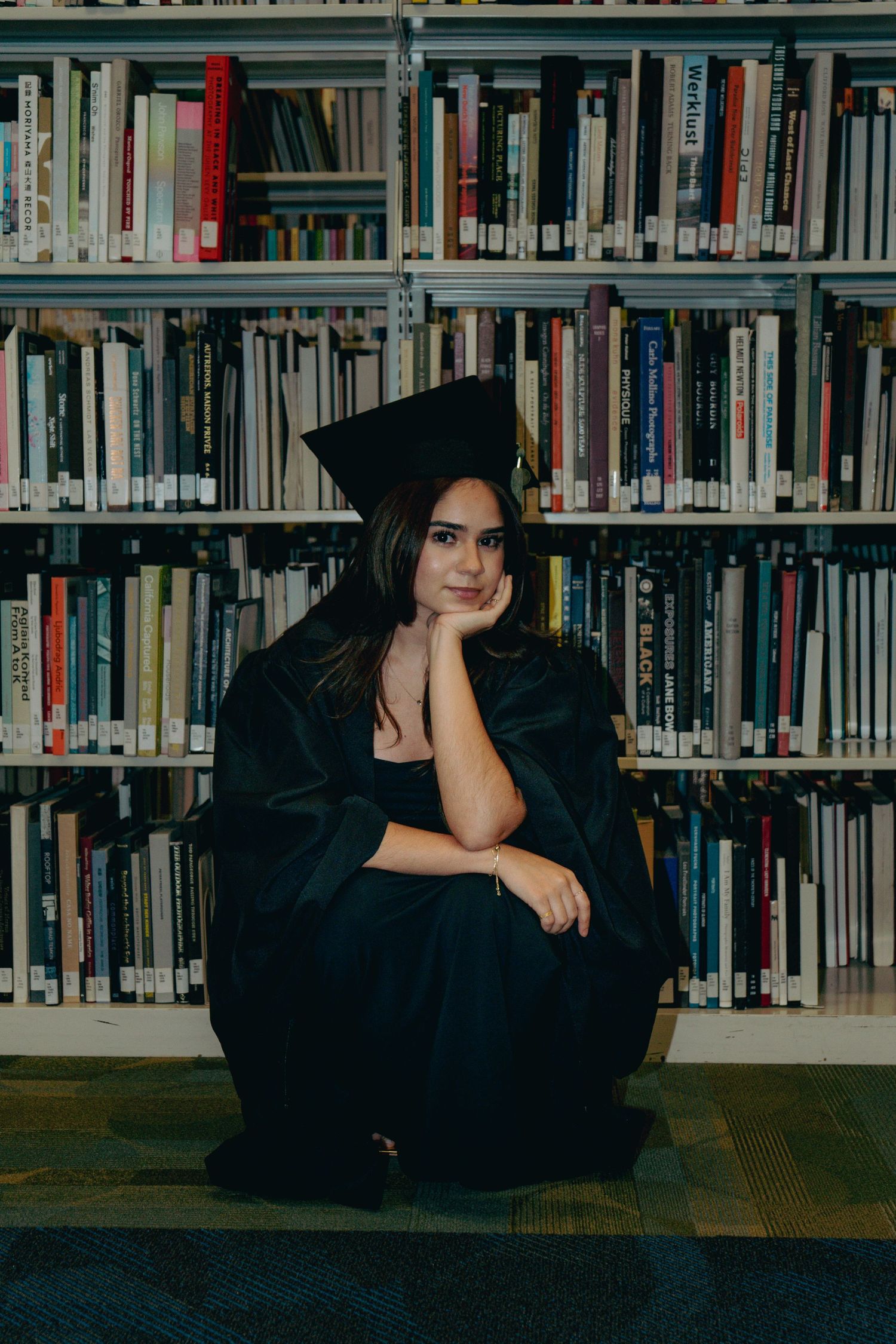 Graduate in cap and gown sitting thoughtfully in front of library bookshelves.