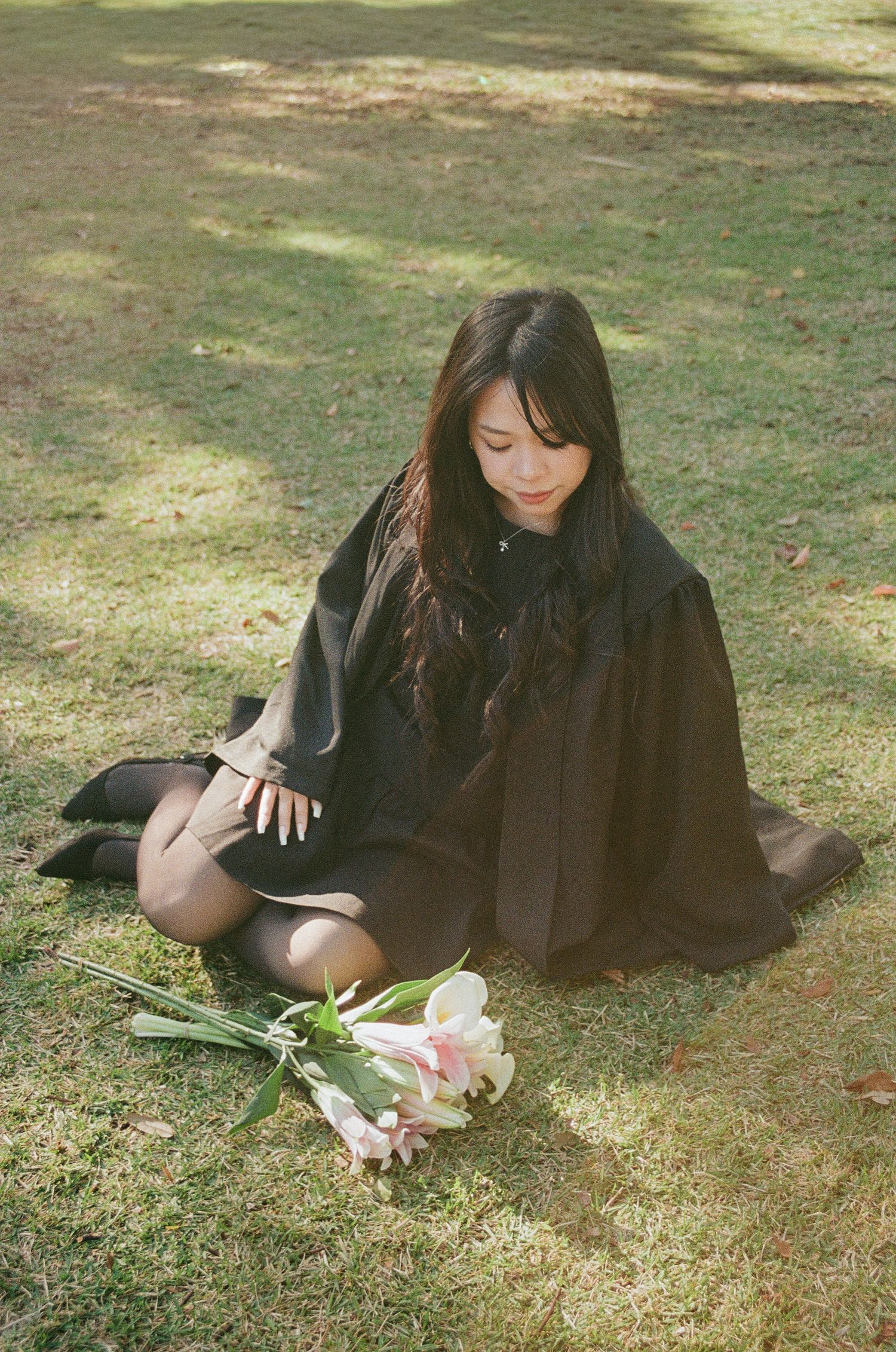 Graduate in black gown sitting on grass with pink and white flowers placed in front of her.
