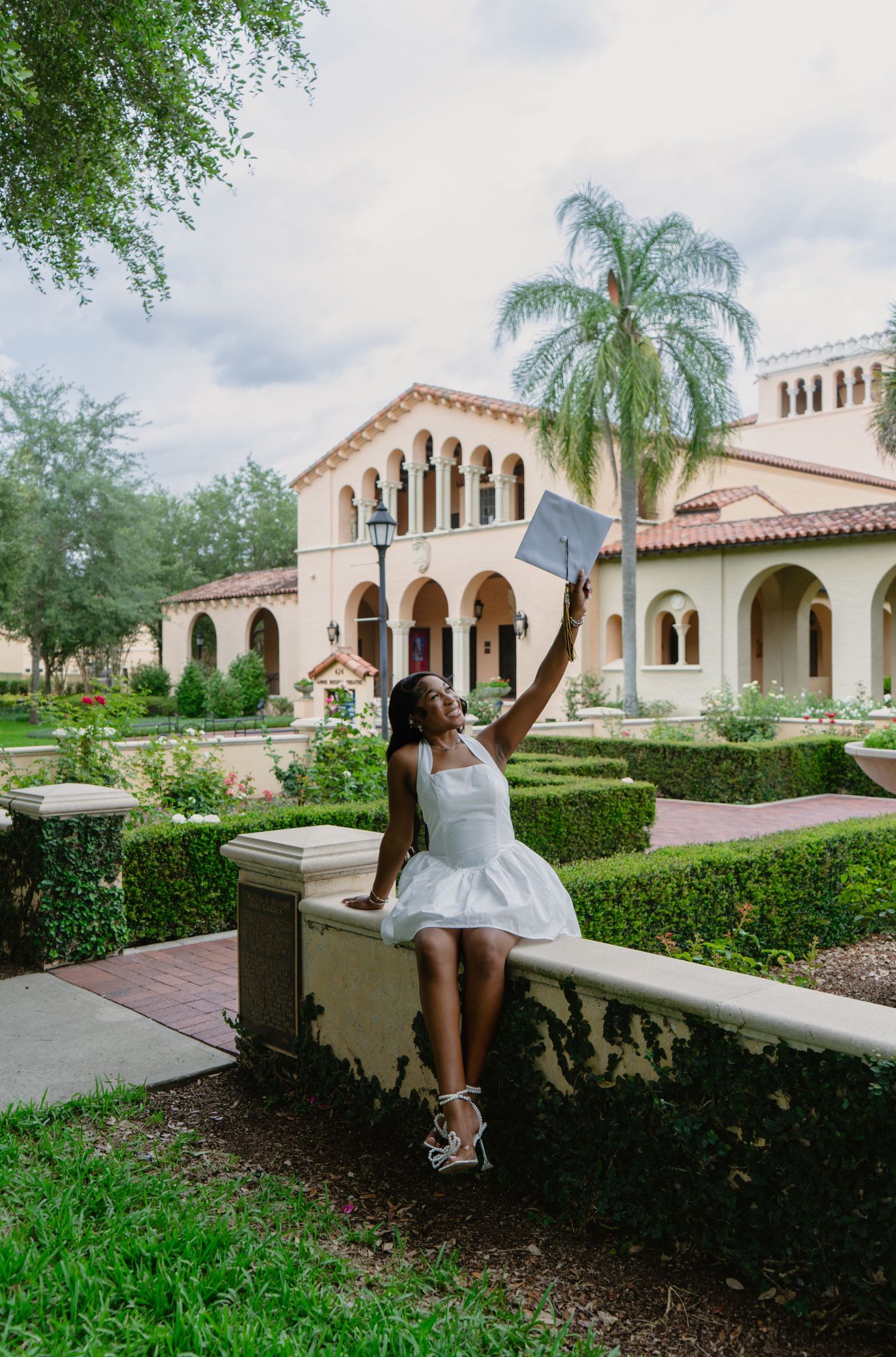 Joyful graduate in white dress raises diploma while sitting on a garden wall at a Florida college campus.
