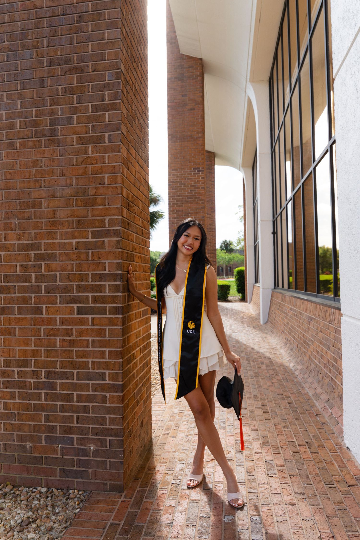 Young woman in graduation attire with black and gold stole holding cap, posing by brick columns.