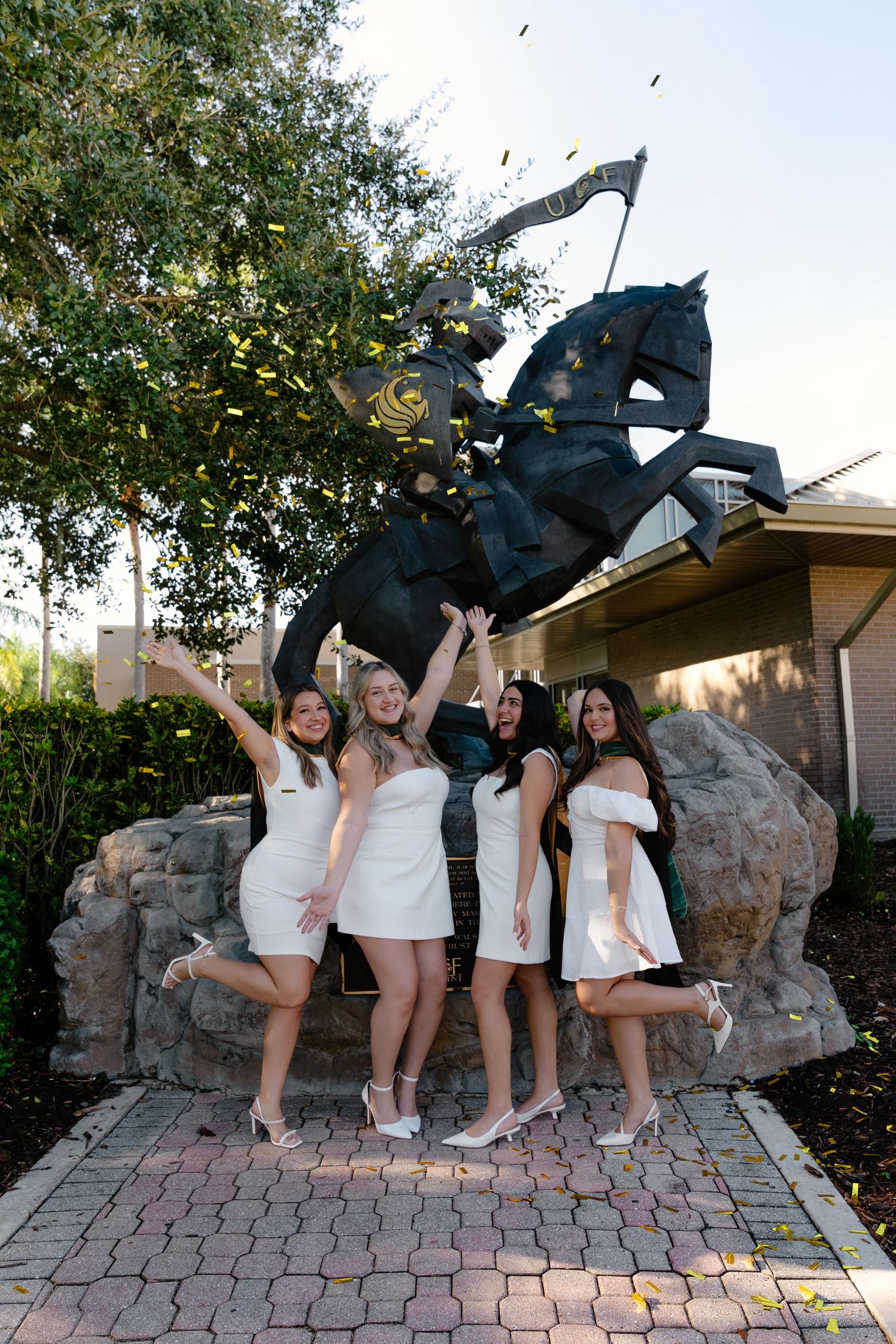 Four women in white dresses celebrating graduation with confetti near a horse statue.