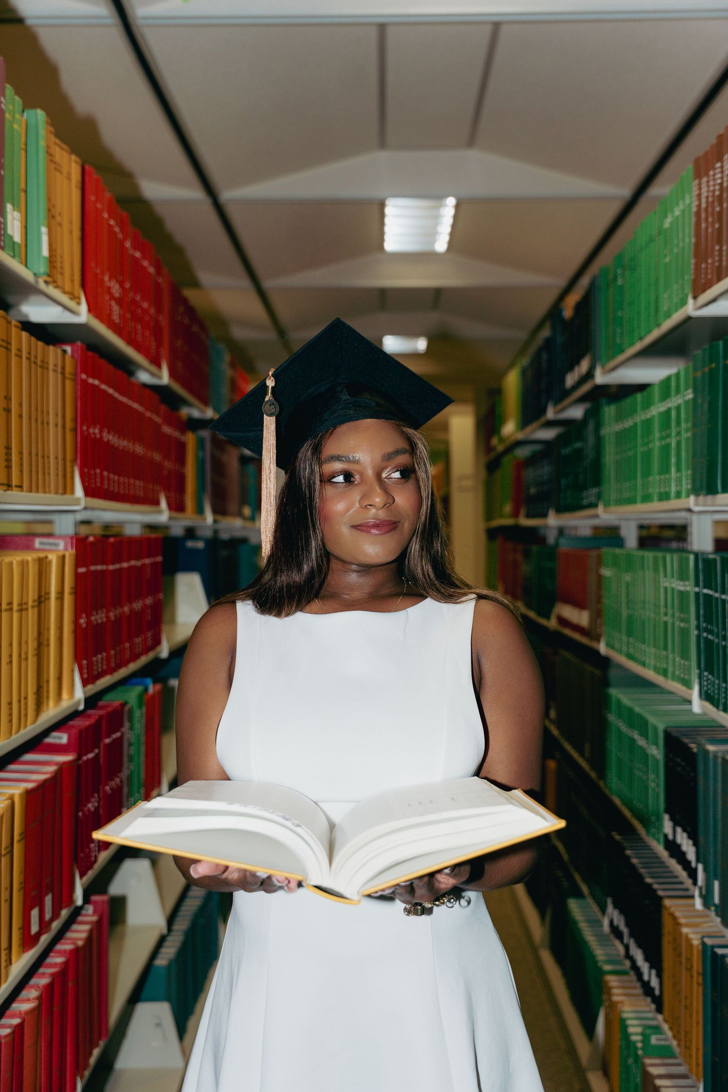 Graduate in white dress and black cap holds open book in colorful university library aisle.