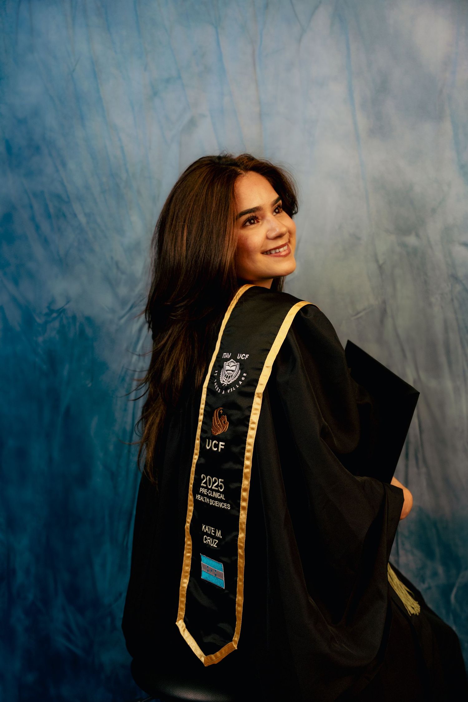Smiling young woman in black graduation gown with UCF 2025 stole posing for professional grad photo.