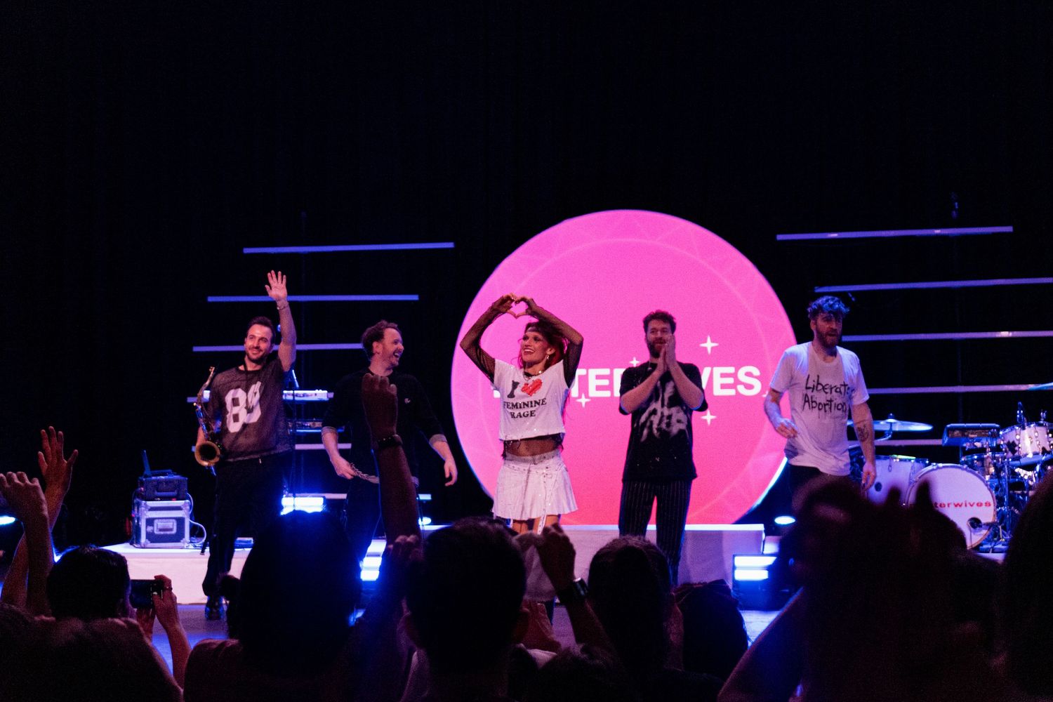 Band performing on stage before pink circular backdrop with enthusiastic crowd.