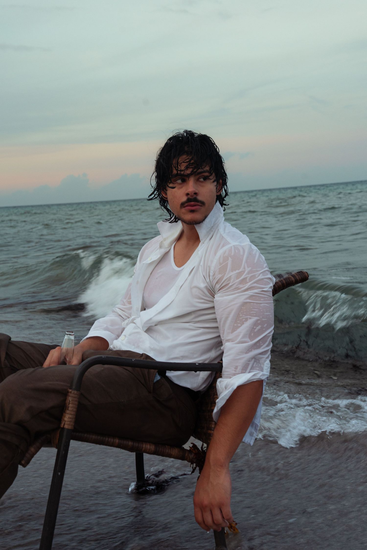 Young man with mustache sitting in chair on beach wearing wet white shirt at dusk.