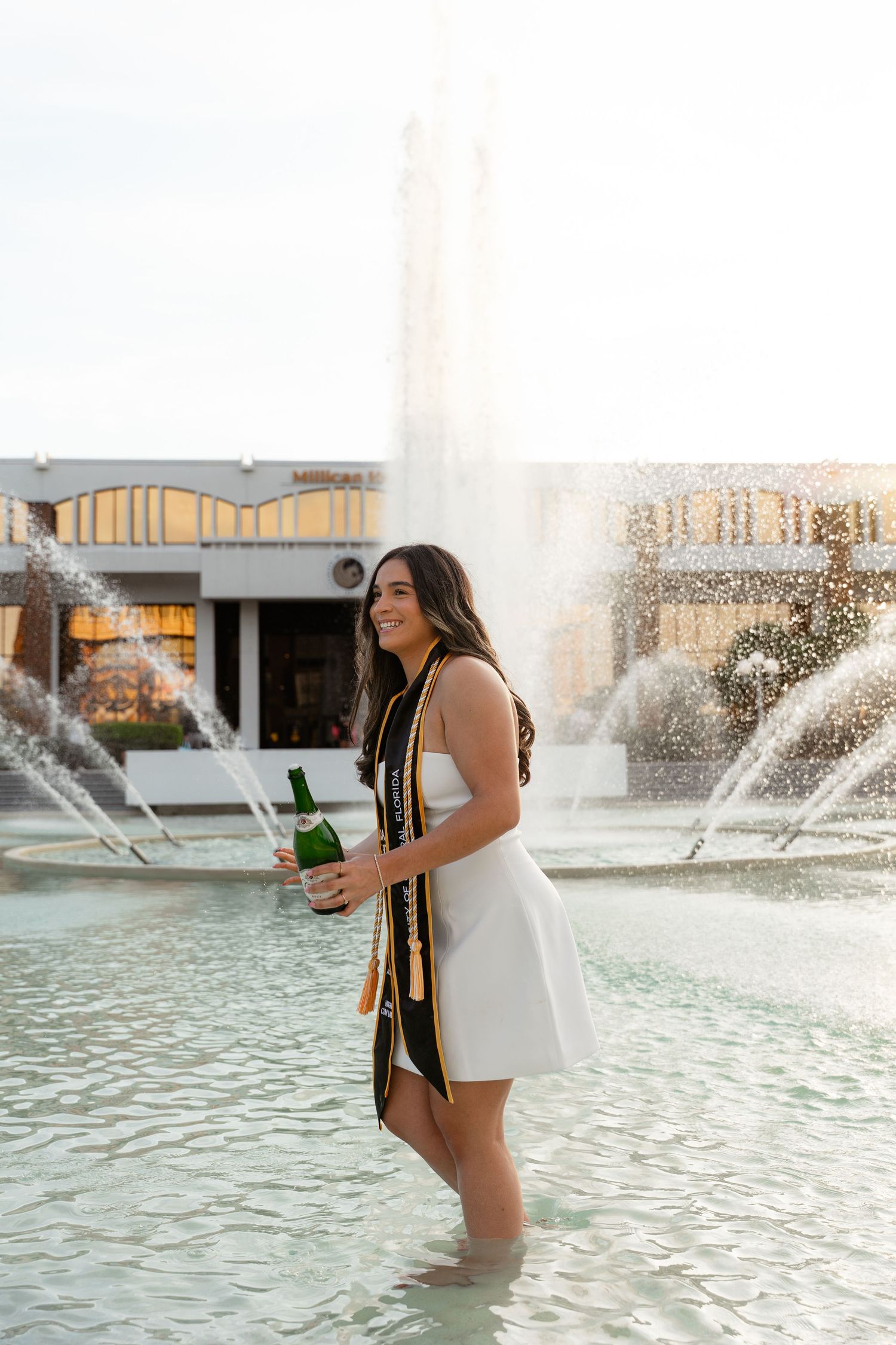 Smiling UCF graduate in white dress and stole holding champagne bottle while standing in a fountain.