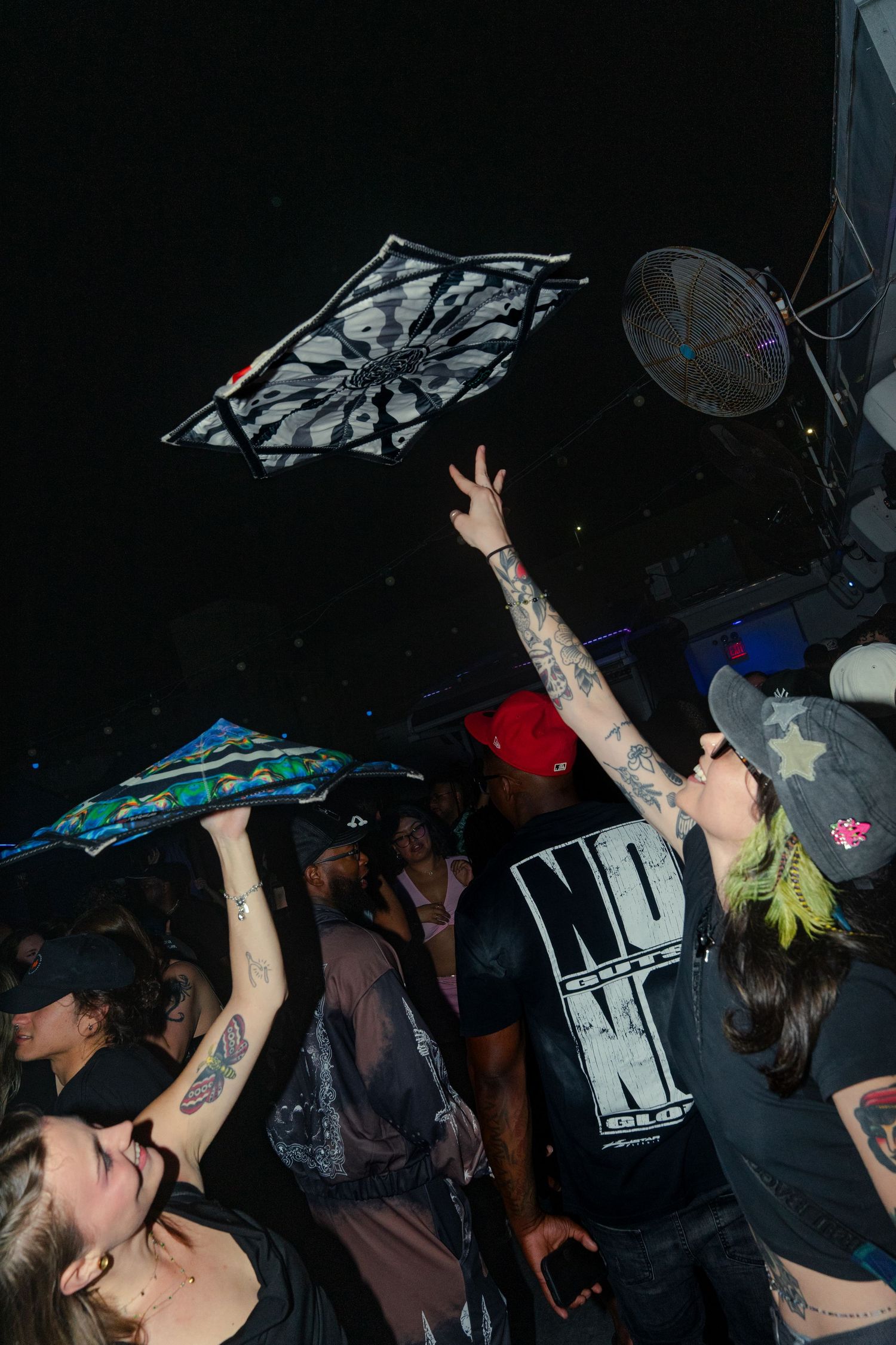 Tattooed woman tosses decorative umbrella at a crowded nightclub event with fans and dark lighting.