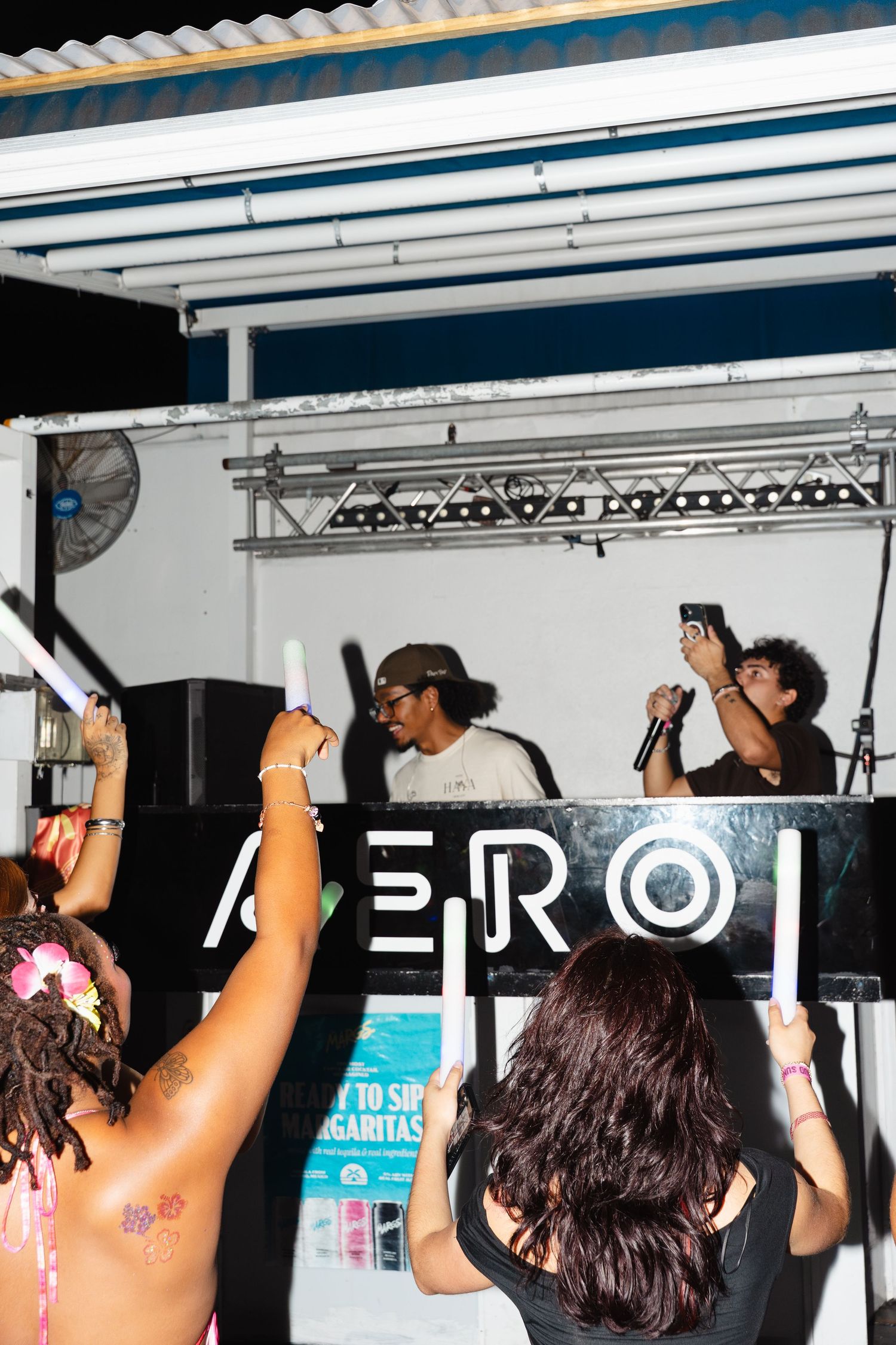 Crowd cheering with glow sticks at an AERO nightclub event with performers on stage holding microphones.