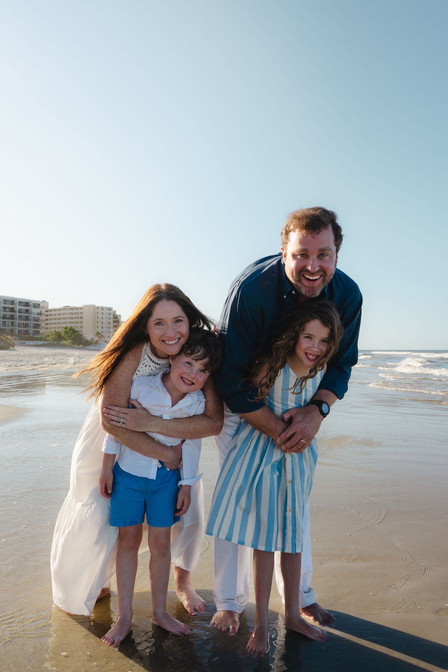 Happy family of four laughing and embracing on a sunny beach with ocean waves in the background.