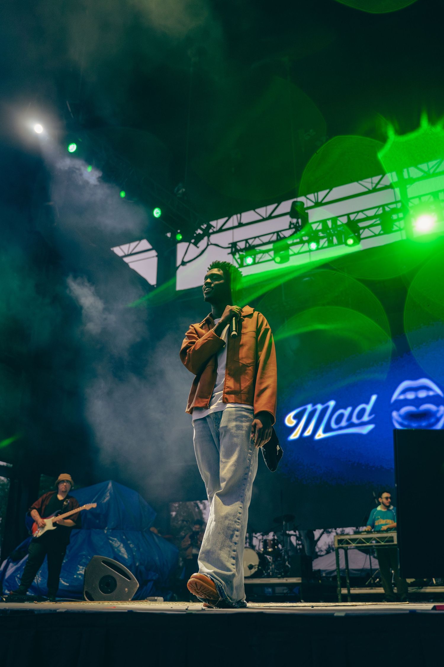 Male rapper performs on a smoky outdoor festival stage under green and blue stage lights.