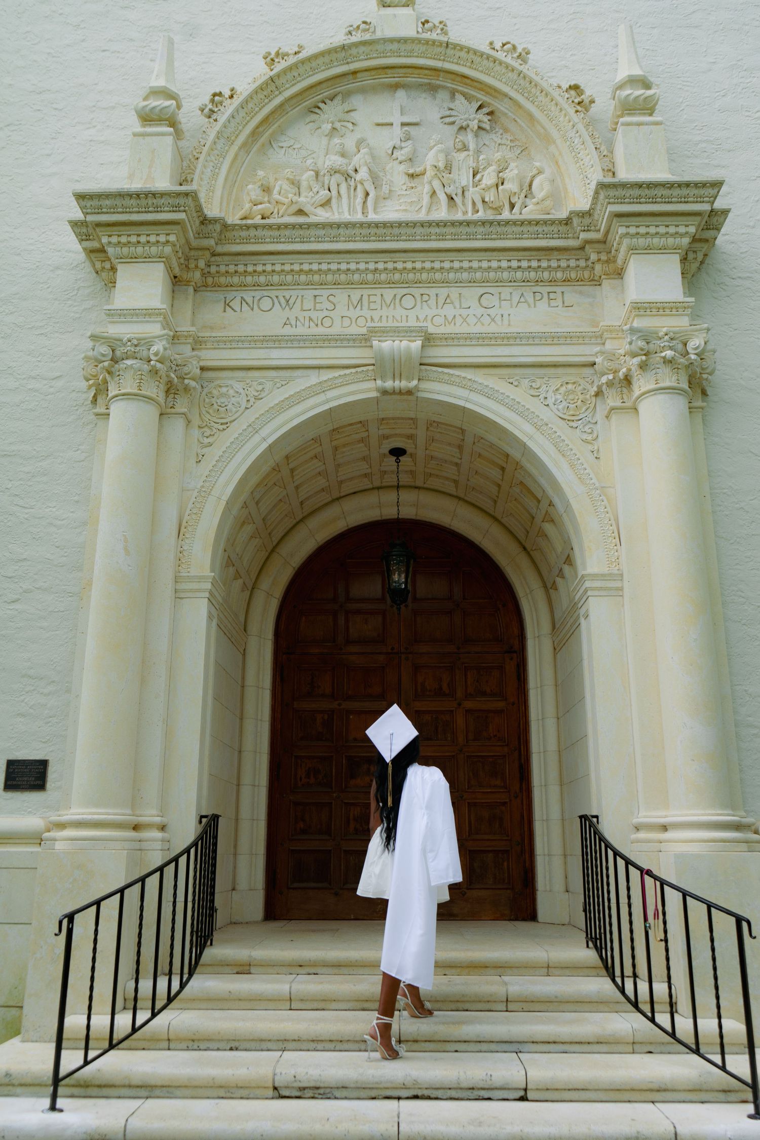 Graduate in white cap and gown stands at the entrance of Knowles Memorial Chapel on a sunny day.