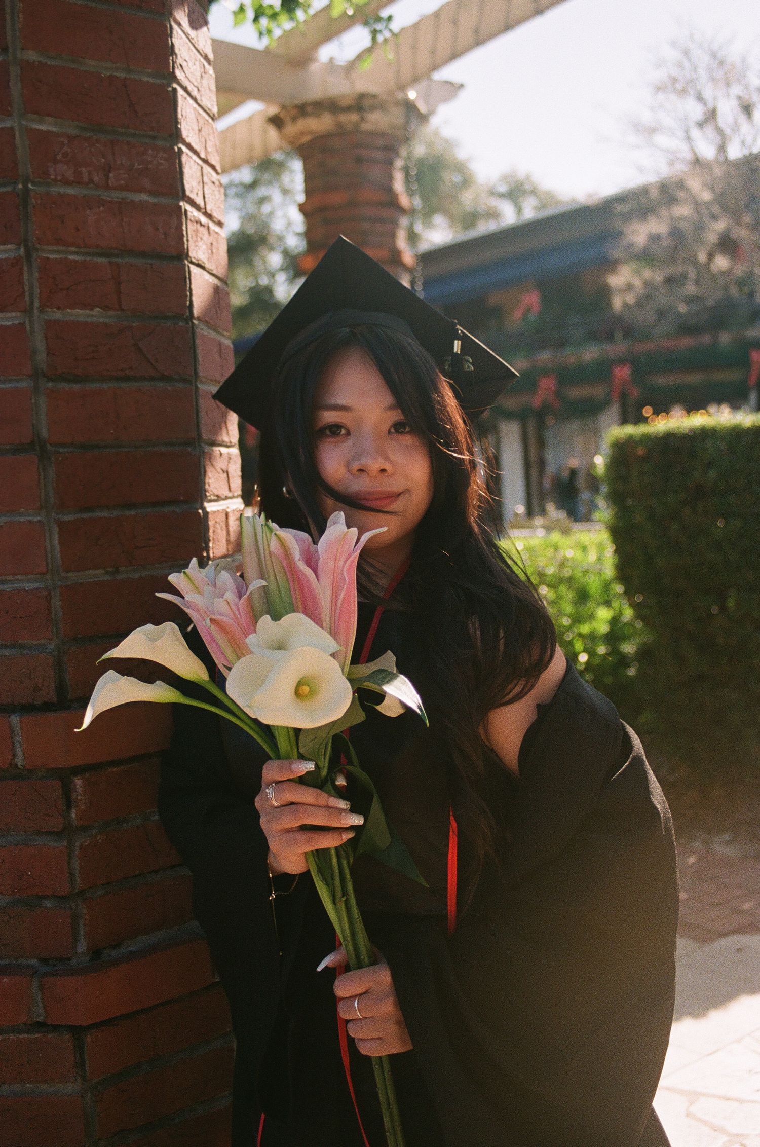 Young woman in graduation cap and gown holding white calla lilies and pink flowers beside a brick column.