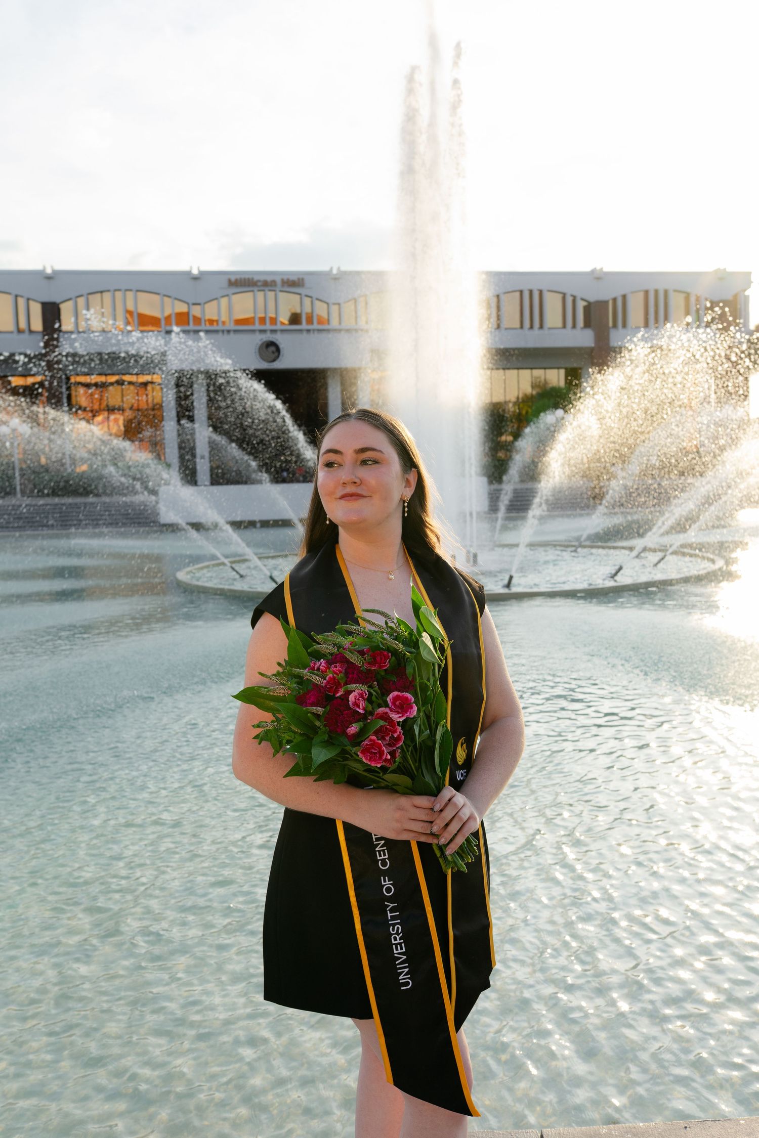 UCF graduate in black gown holding red roses in front of a campus fountain.