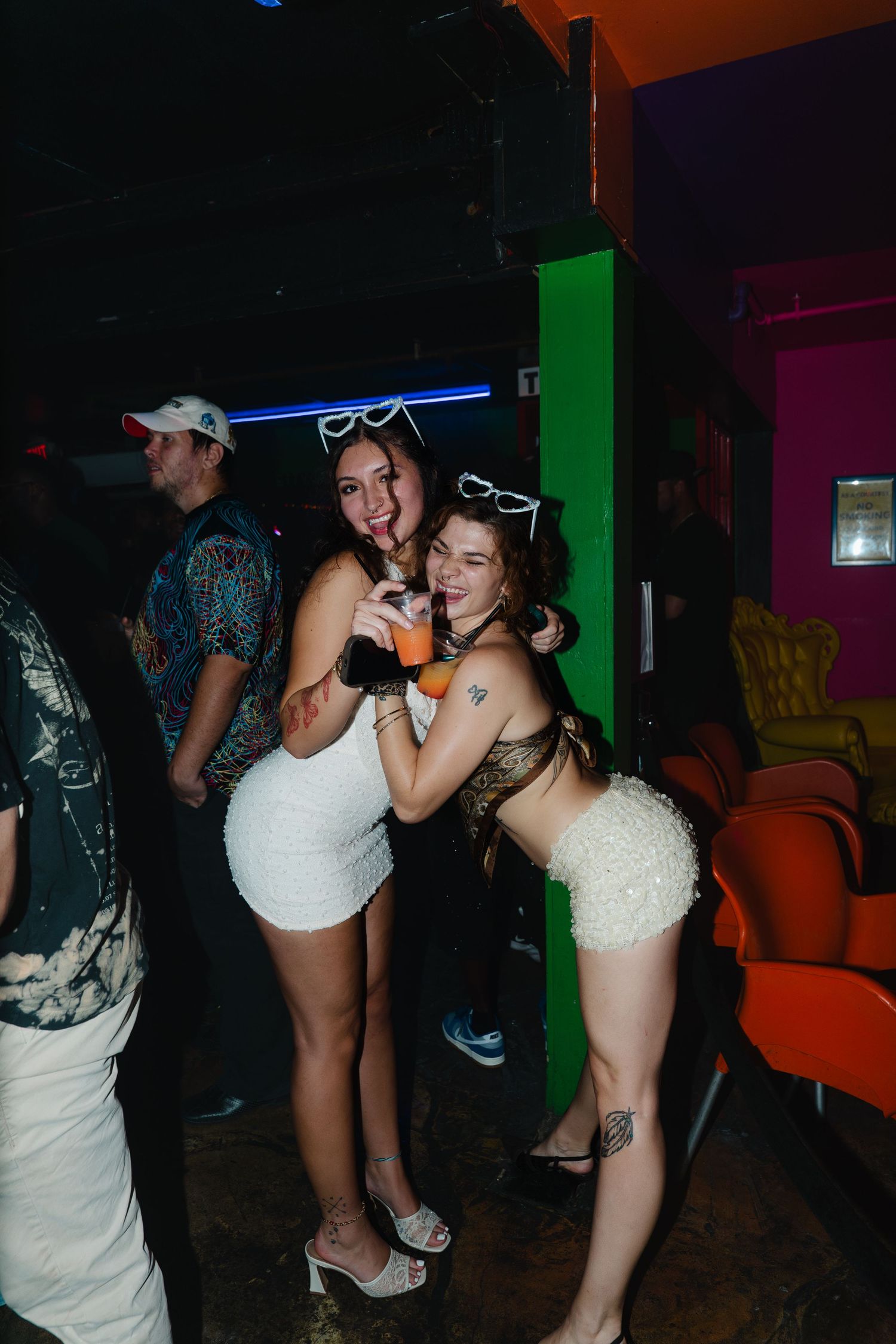 Two young women in white outfits posing together at a nightclub party, smiling and holding drinks.