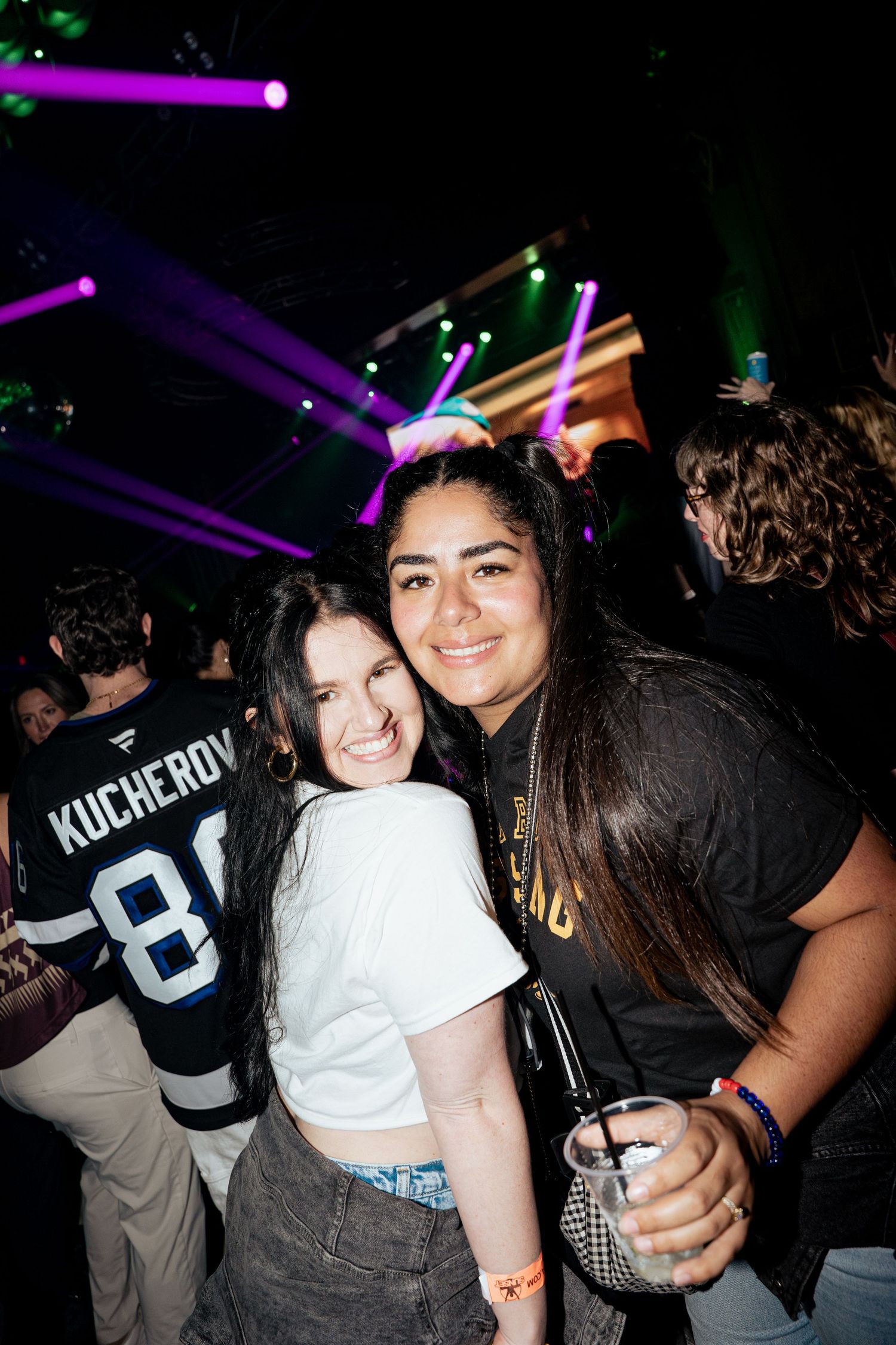 Two young women smiling and posing together at a nightclub with colorful stage lights in the background.