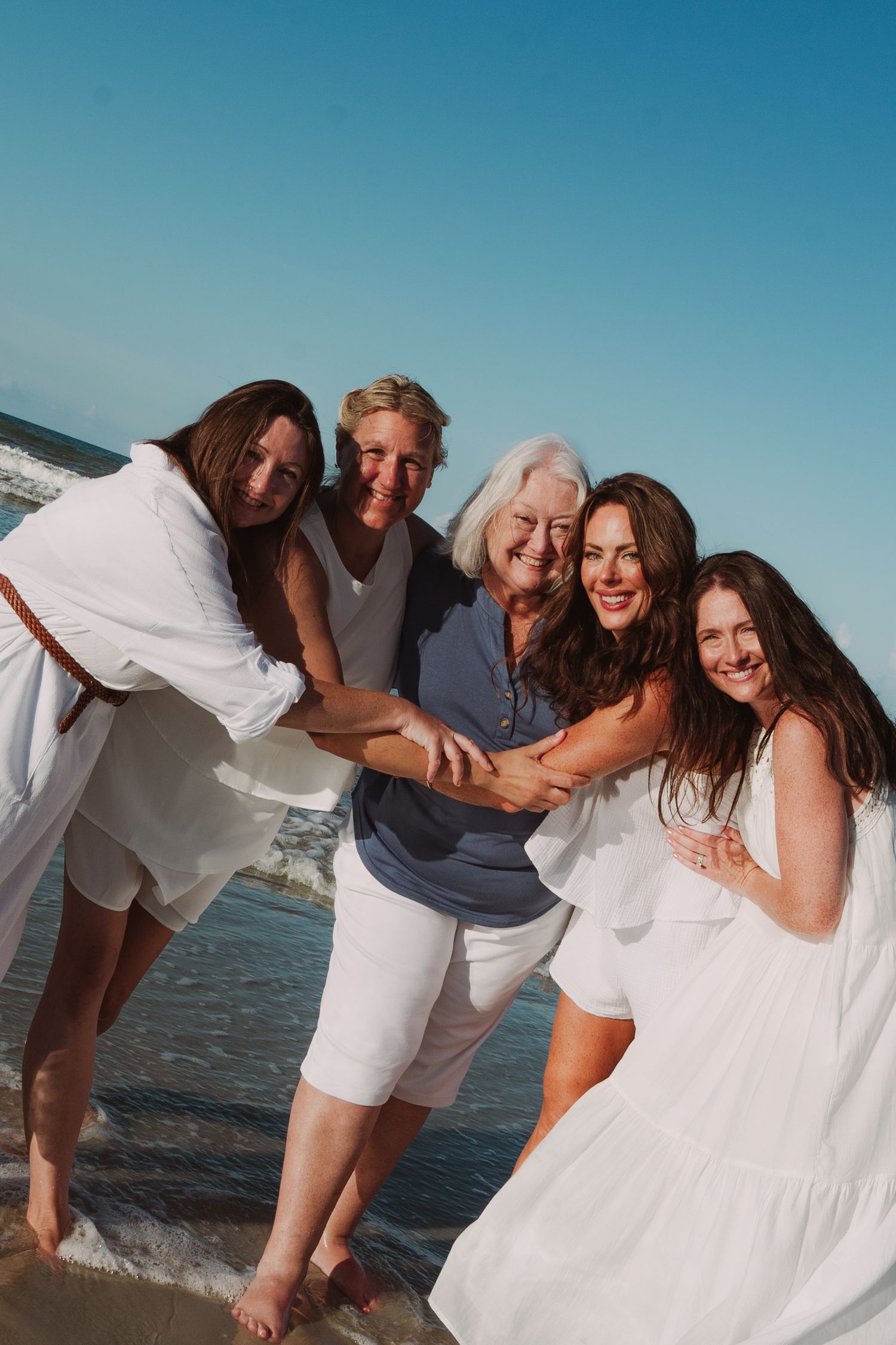 Five women of different generations hugging and smiling together on sunny beach in white outfits.
