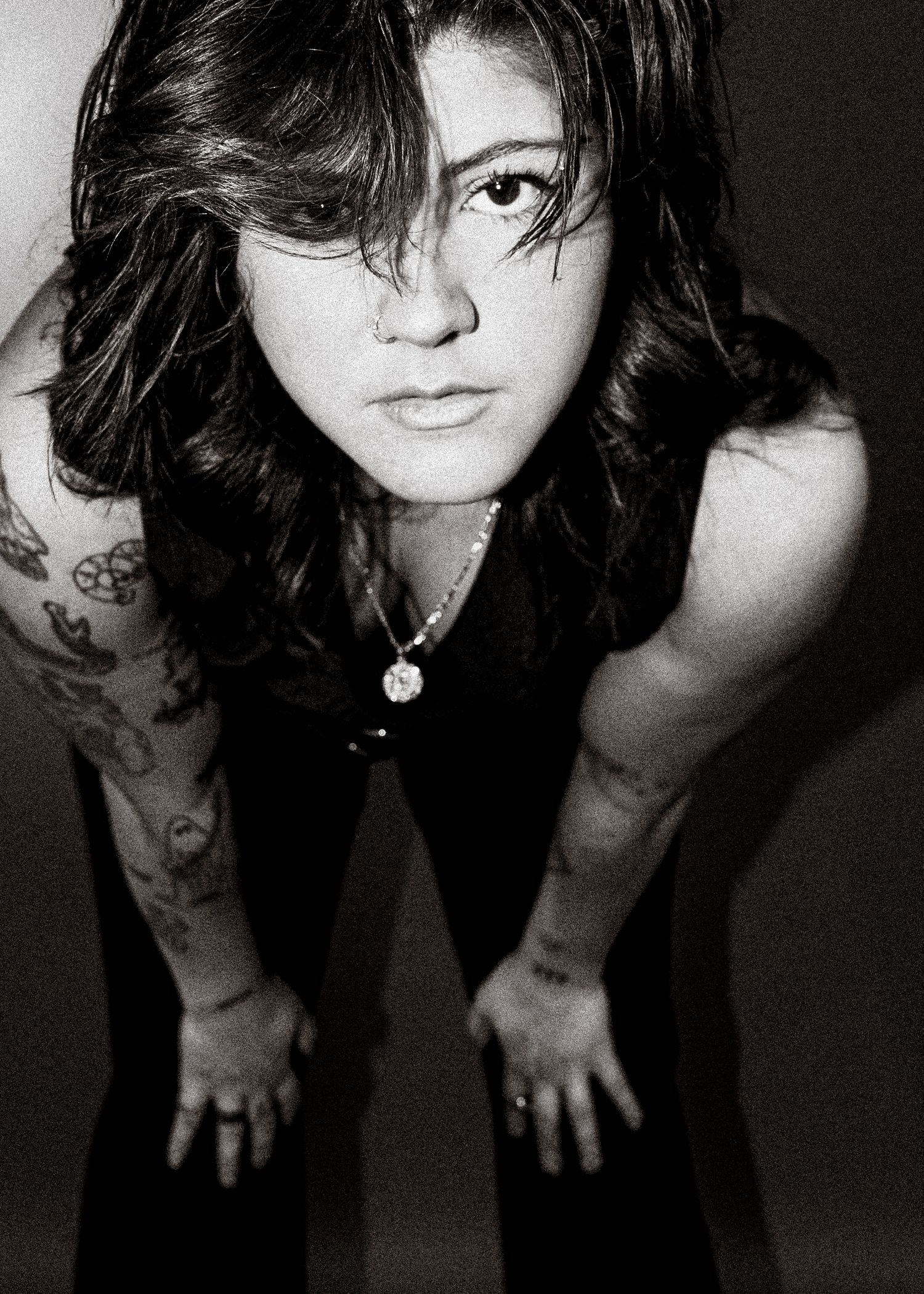 Black and white portrait of tattooed woman with dark hair leaning forward toward camera intensely.