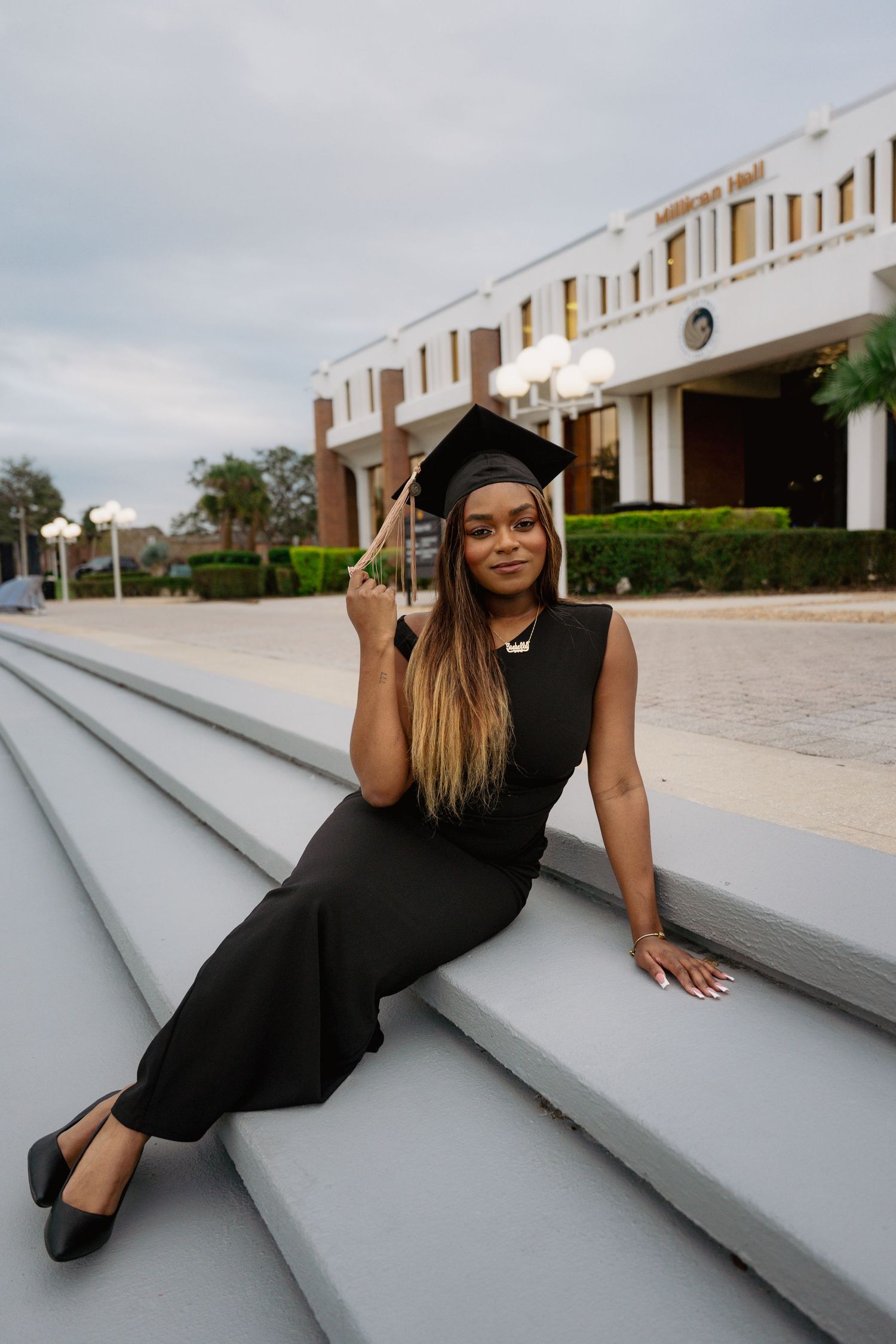 Female graduate in black outfit and cap sitting on outdoor steps in front of university building at dusk.