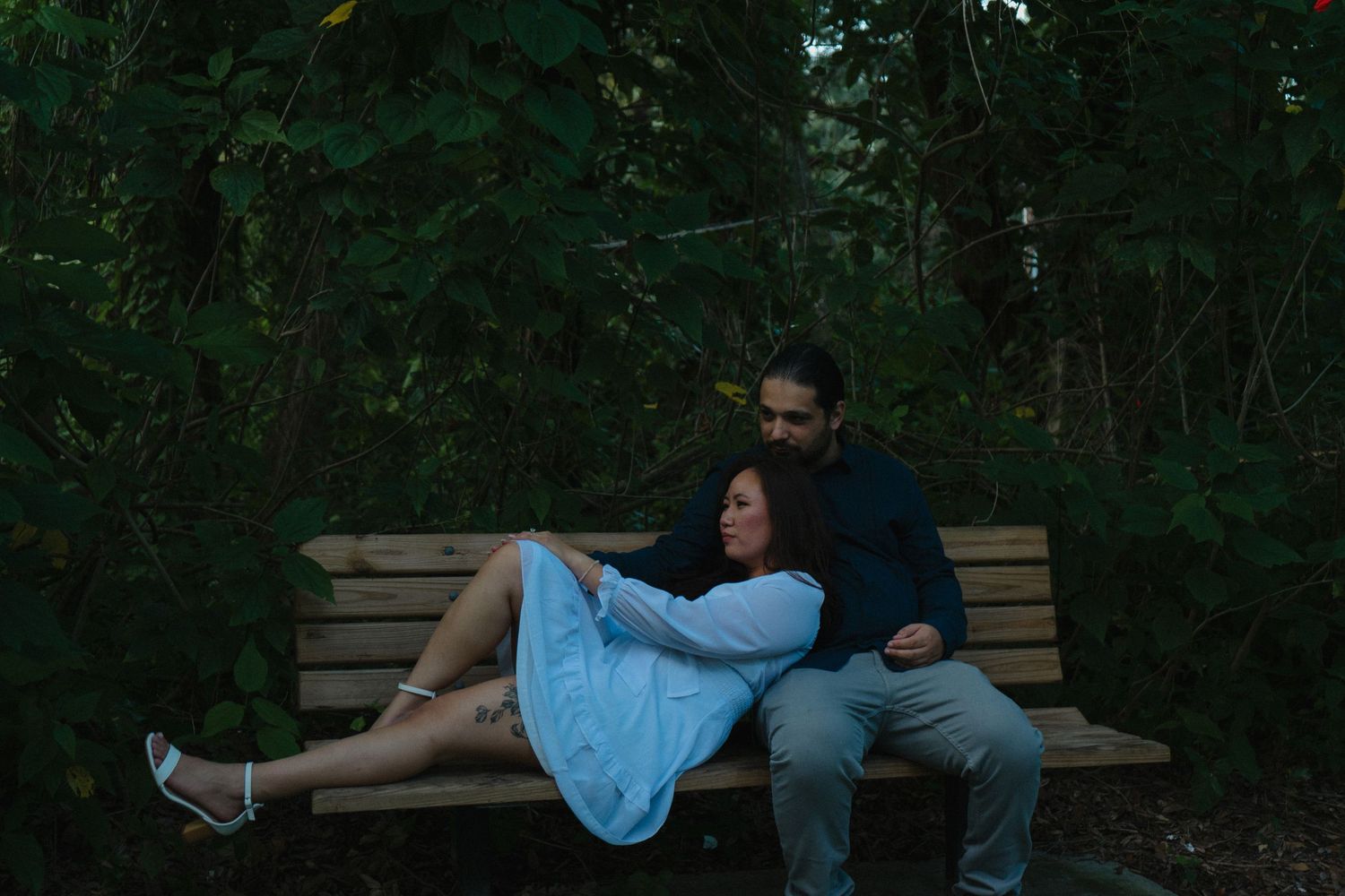 Couple sitting closely on a wooden bench surrounded by dark green forest foliage.