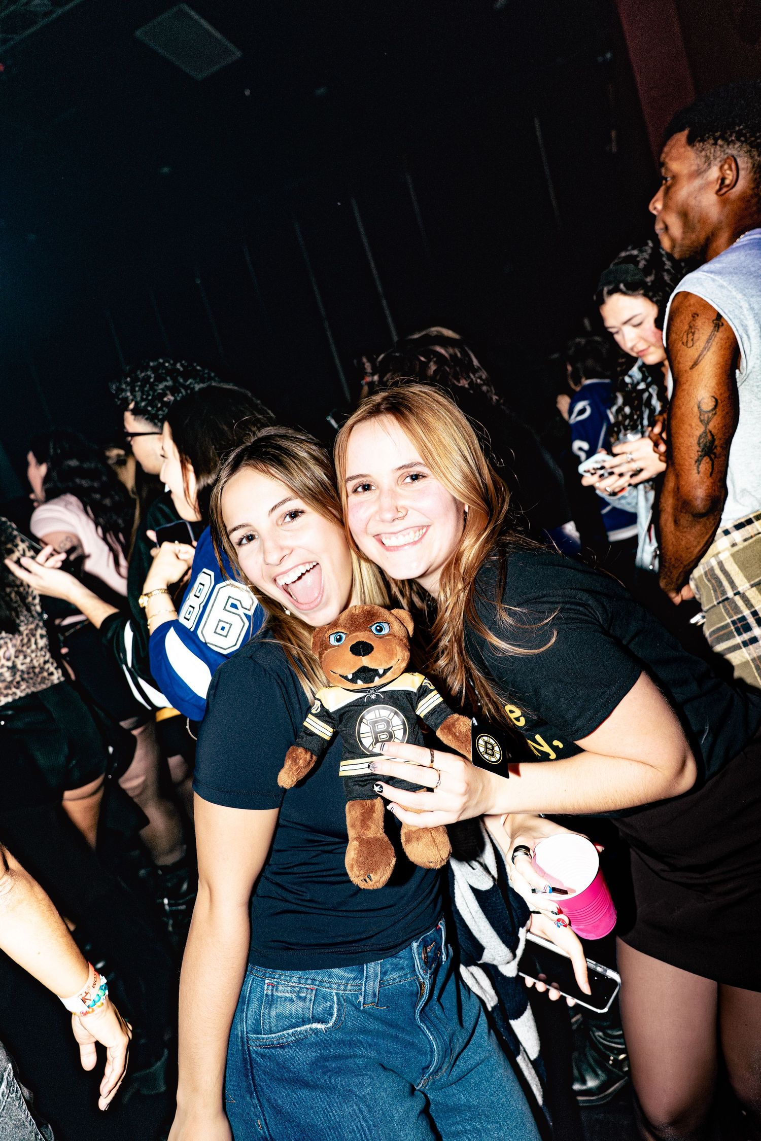 Two smiling women pose with a teddy bear at a lively nightclub event.