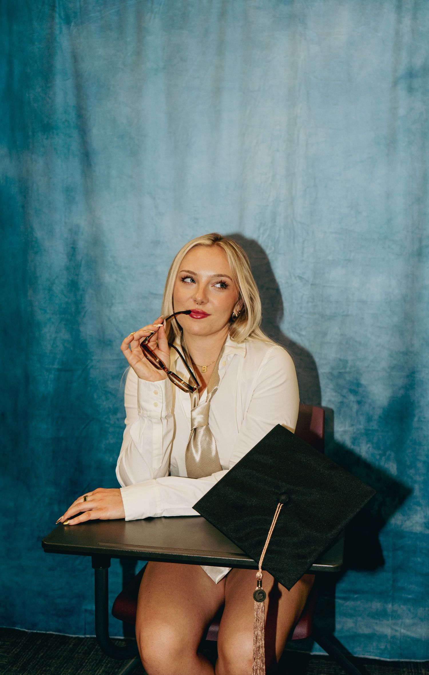 Blonde graduate in white blazer sits at school desk holding glasses beside graduation cap on blue backdrop.