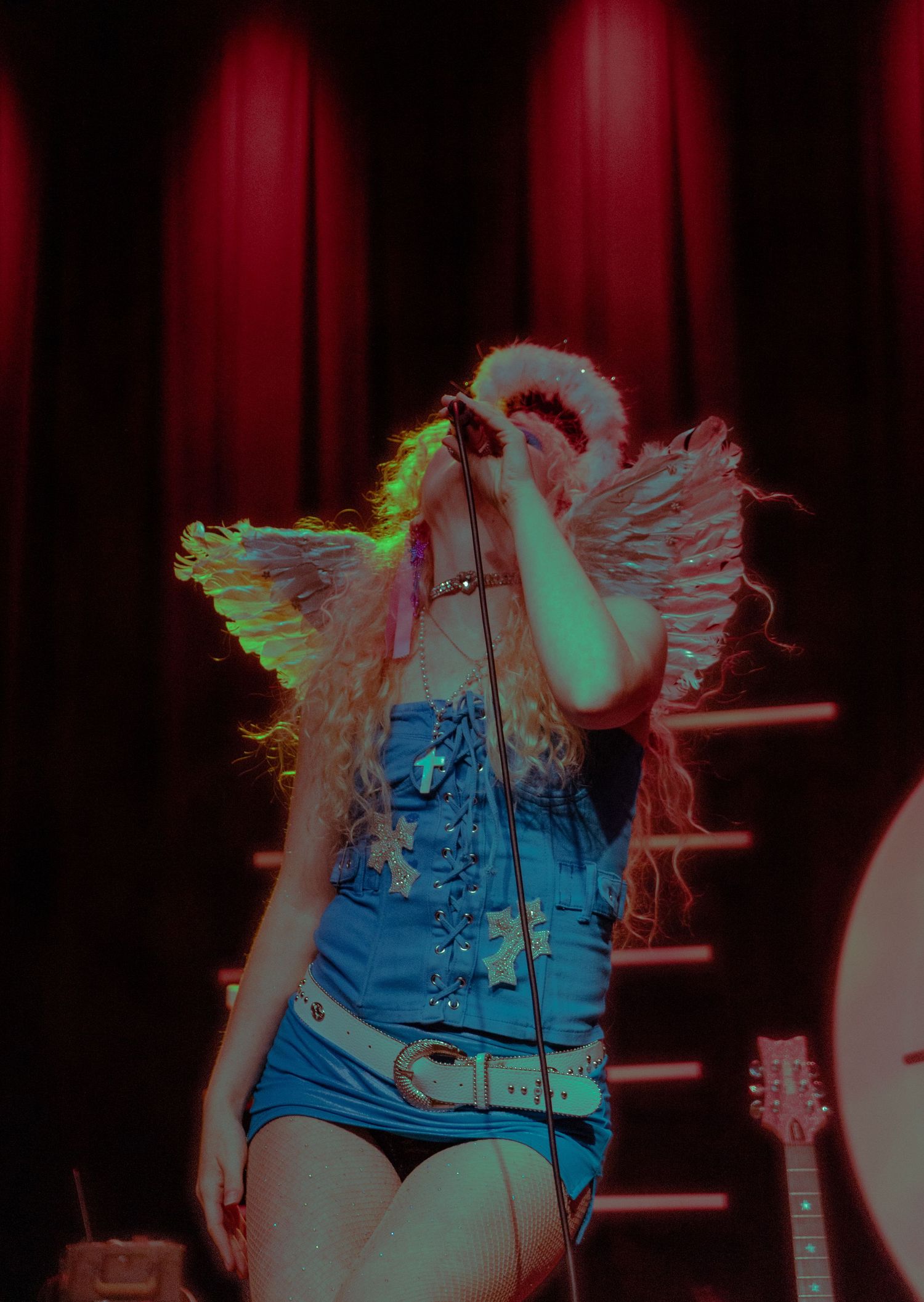 Female performer in denim corset with angel wings raises microphone under red and green stage lighting.