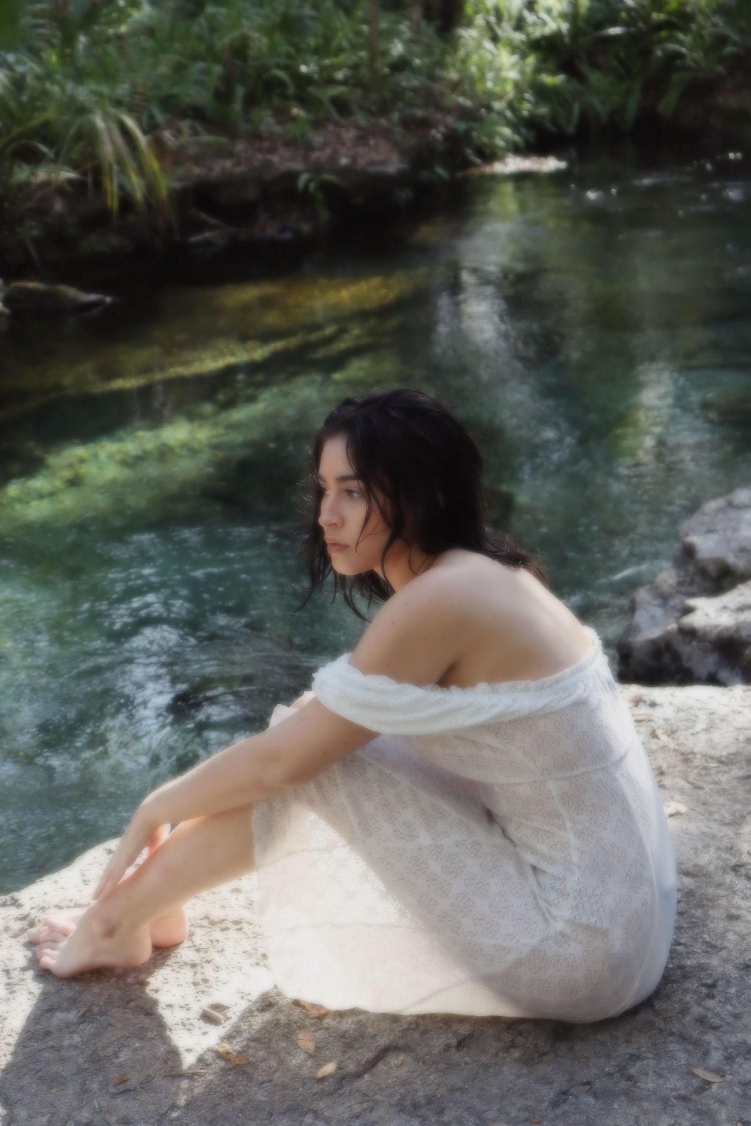 Young woman in white off-shoulder dress sitting on rocks beside a serene green river.
